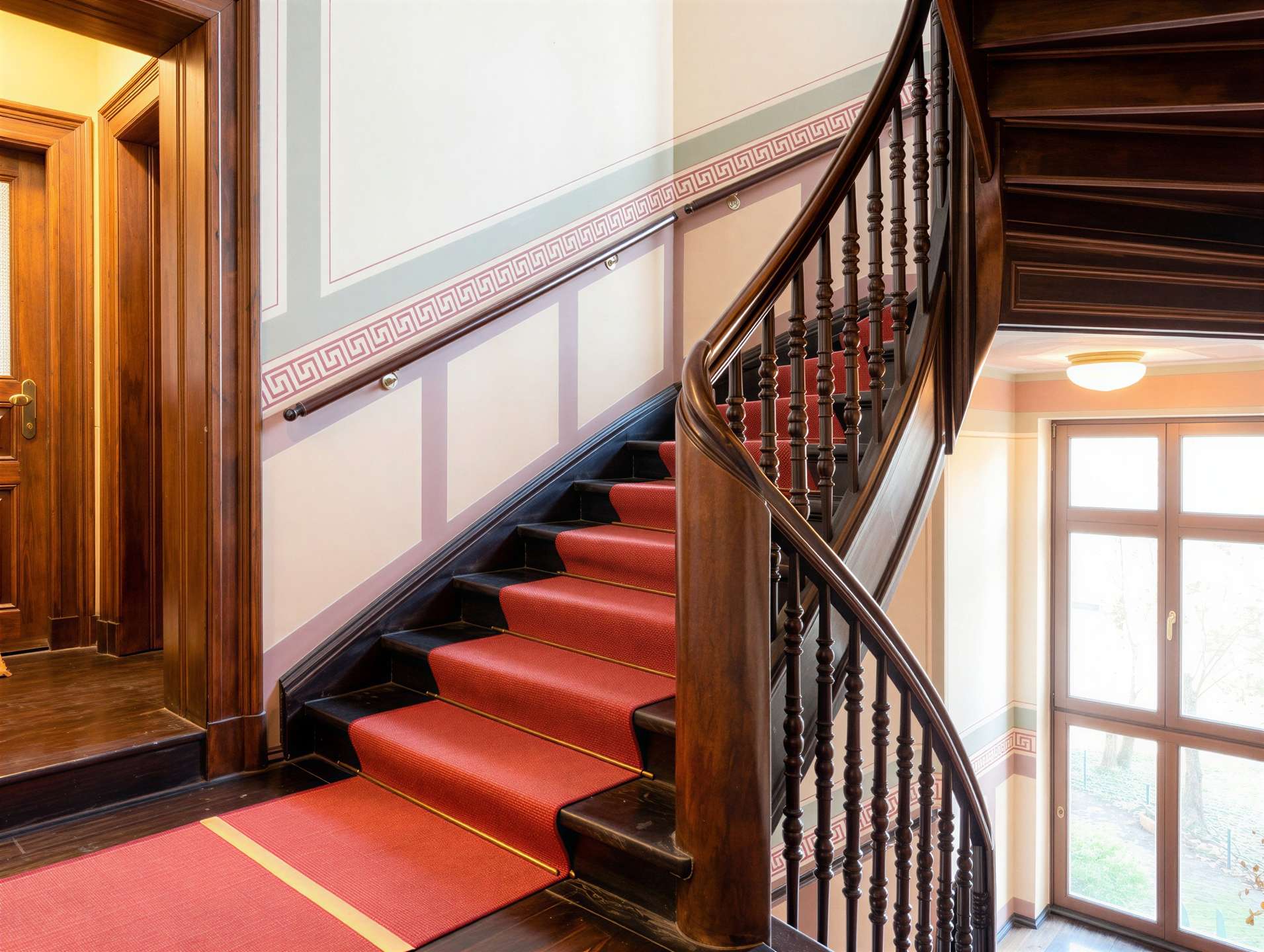 Curved wooden staircase with a red carpet, ornate balusters, and panelled walls in a sunlit foyer.