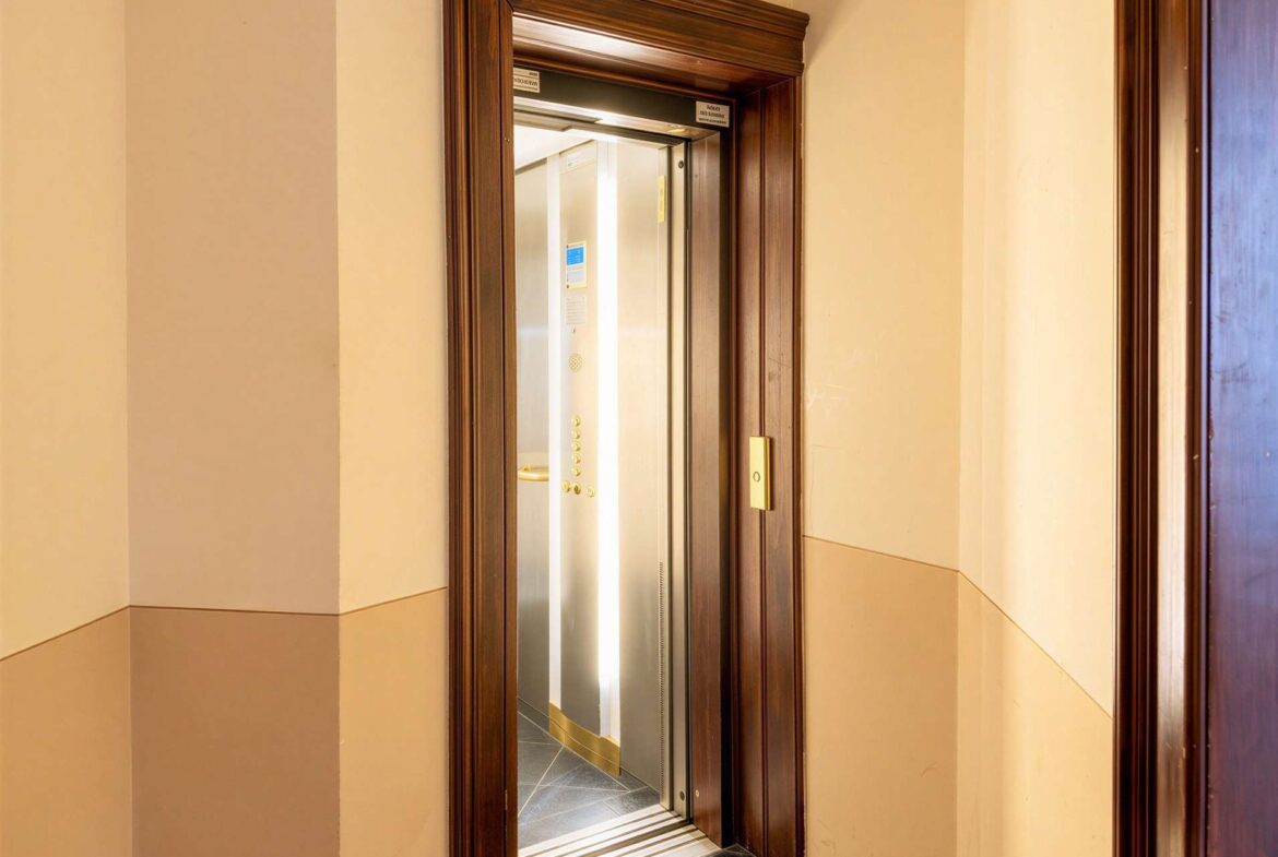 Open elevator with stainless steel doors in a dark wood frame, beige walls and tiled floor beyond the doorway.