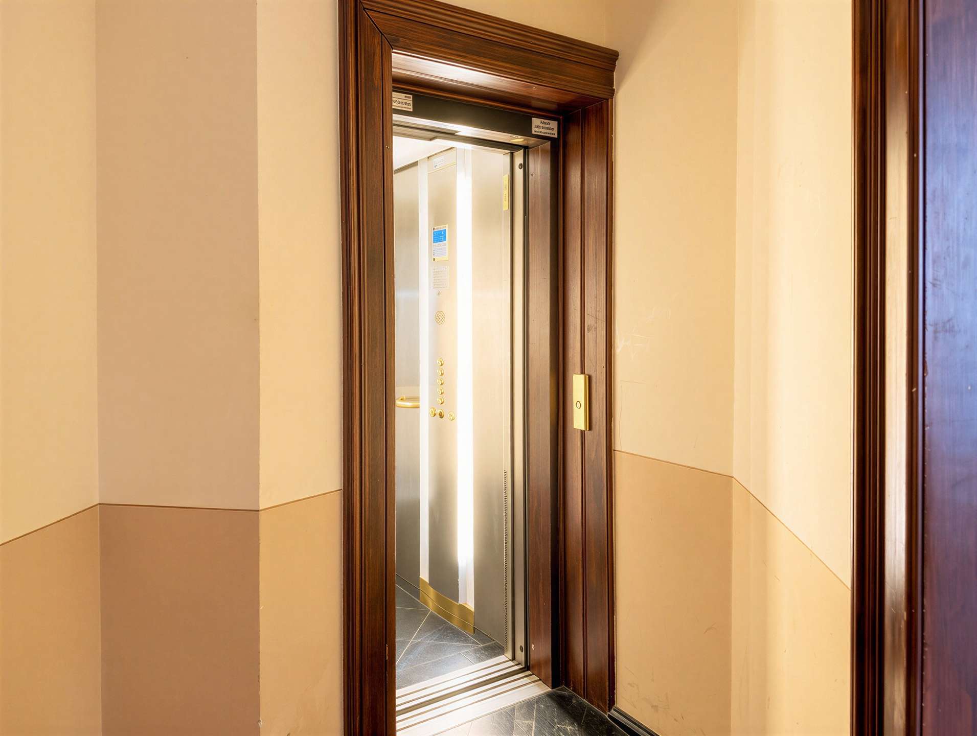 Open elevator with stainless steel doors in a dark wood frame, beige walls and tiled floor beyond the doorway.