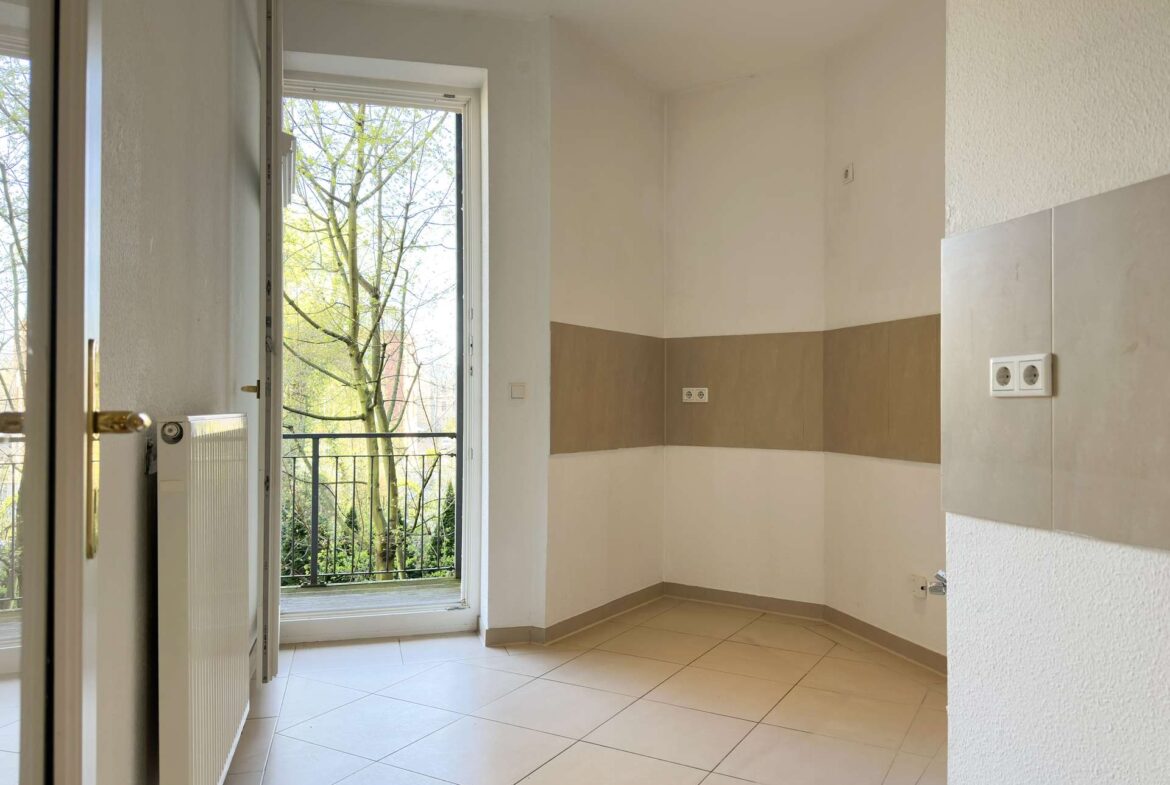 Empty kitchen corner with tiled floor, beige wall tiling, radiator to the left, and an open door leading to a balcony with trees outside.