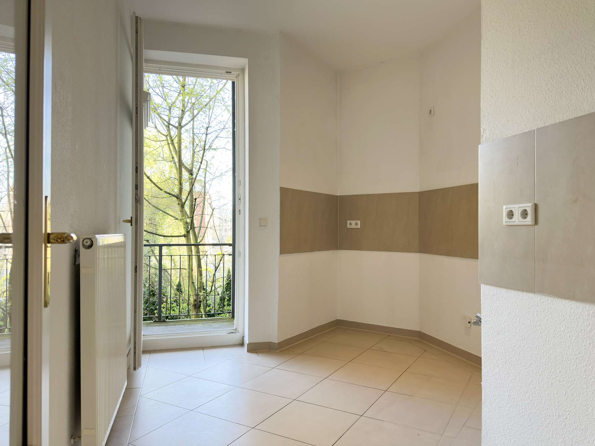 Empty kitchen corner with tiled floor, beige wall tiling, radiator to the left, and an open door leading to a balcony with trees outside.