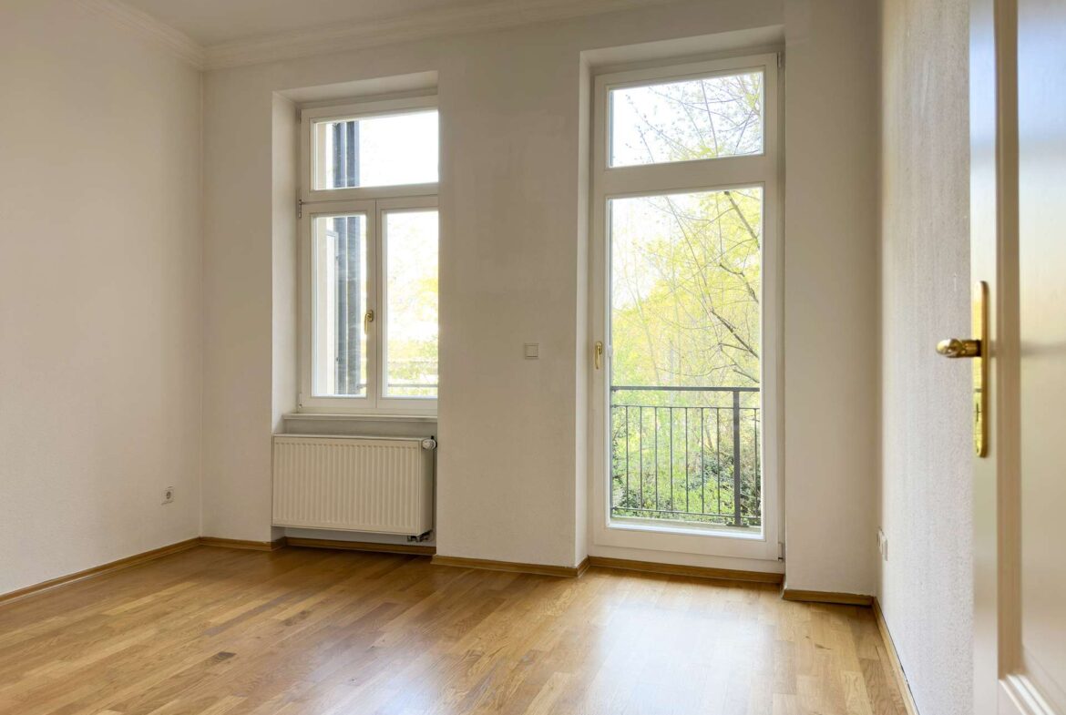 Bright empty room with light wooden floor, white walls, and a tall window overlooking a balcony and trees outside; radiator beneath window.