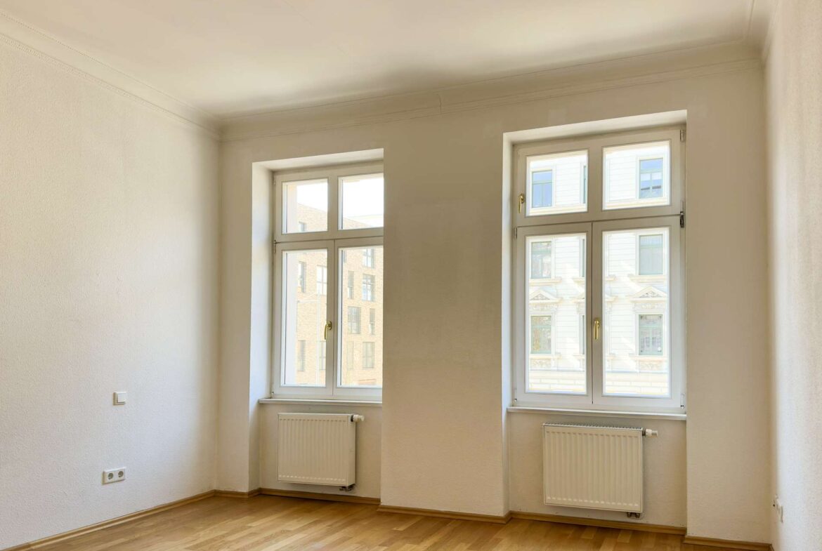 Empty, sunlit room with light wood flooring, white walls, and two large white-framed windows with radiators beneath.