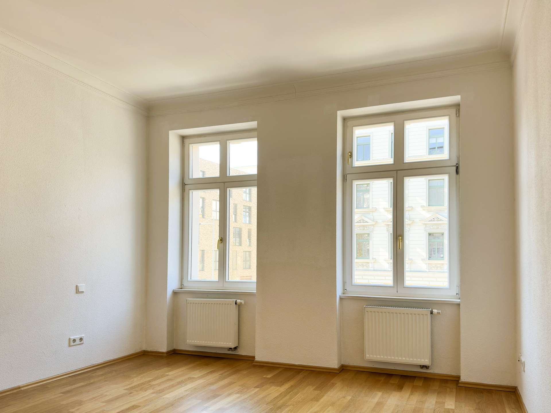 Empty, sunlit room with light wood flooring, white walls, and two large white-framed windows with radiators beneath.