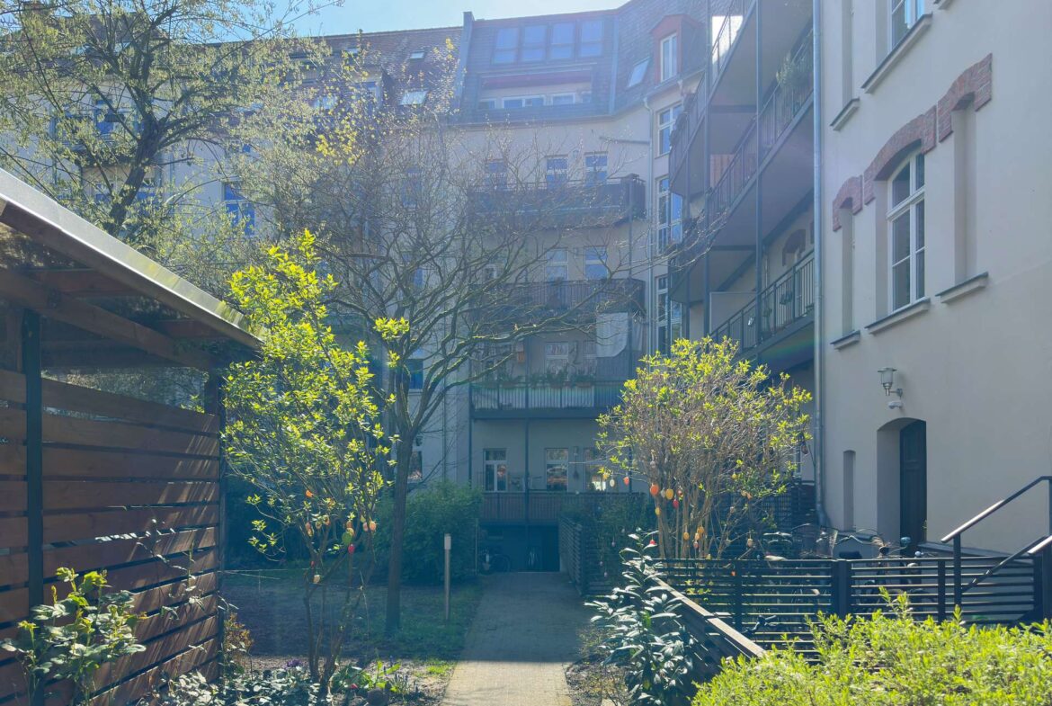 Sunlit apartment courtyard with a paved walkway, trees, shrubs, and balconies along tall building facades on both sides.