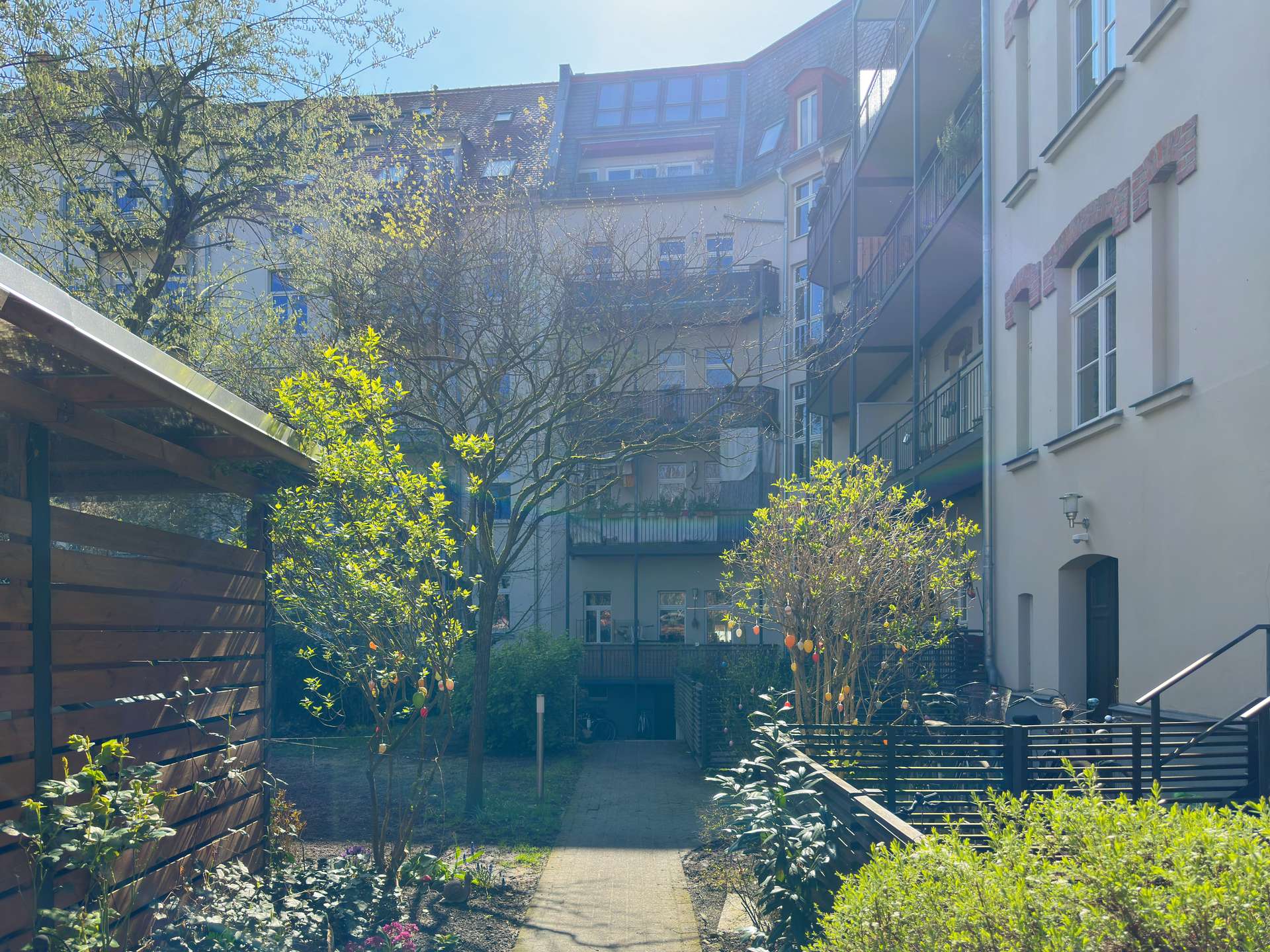 Sunlit apartment courtyard with a paved walkway, trees, shrubs, and balconies along tall building facades on both sides.