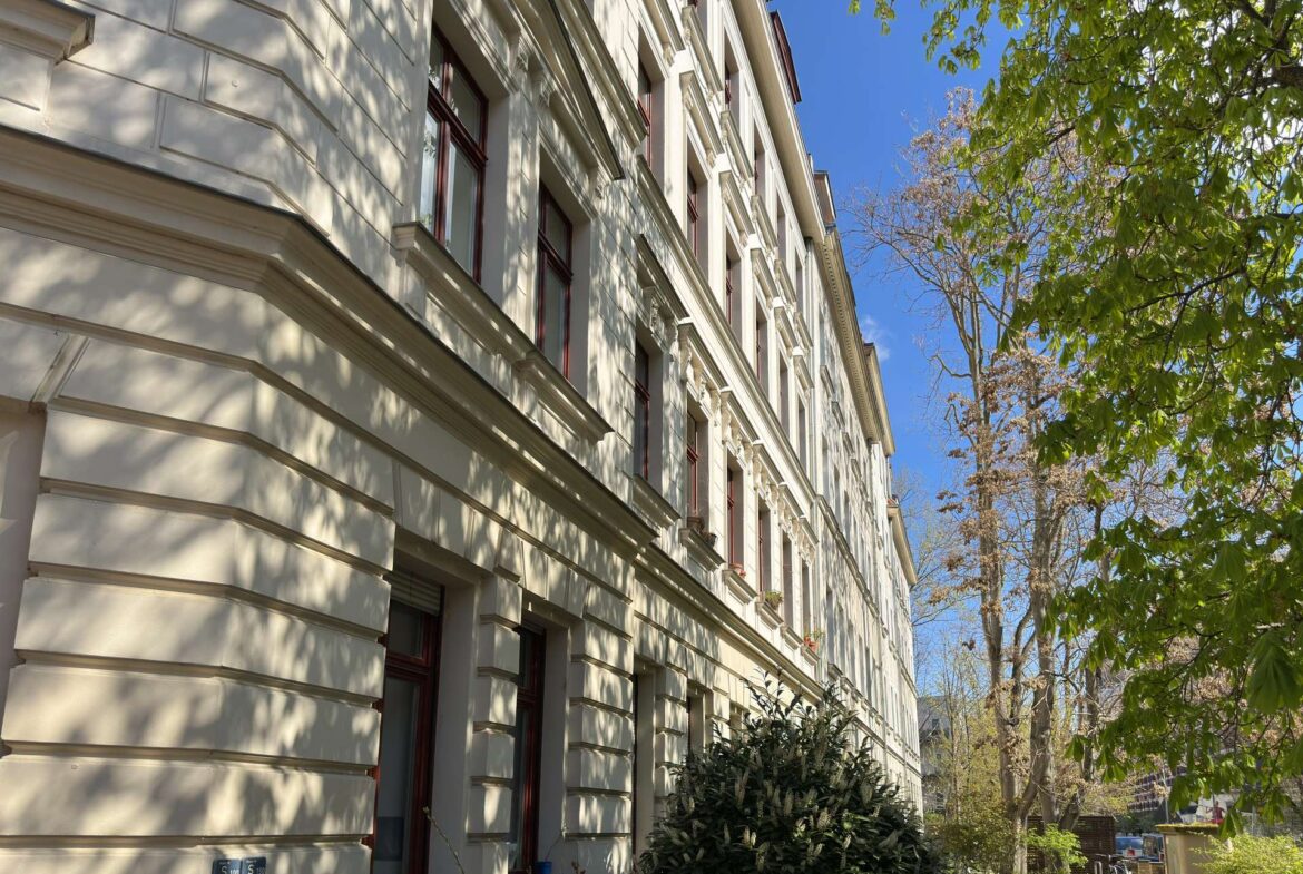 Facade of a light-colored apartment building with ornate cornices and red-framed windows under a sunny blue sky with tree shadows on the wall.
