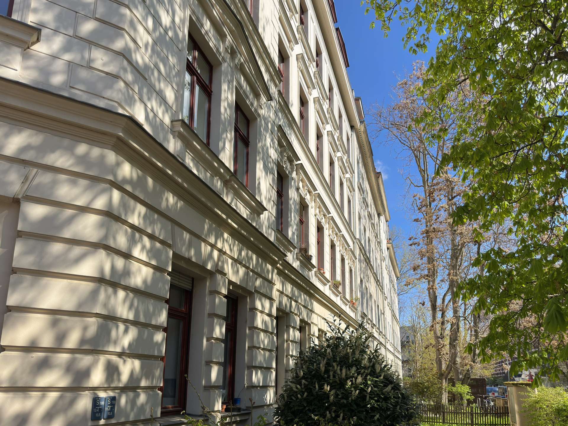 Facade of a light-colored apartment building with ornate cornices and red-framed windows under a sunny blue sky with tree shadows on the wall.