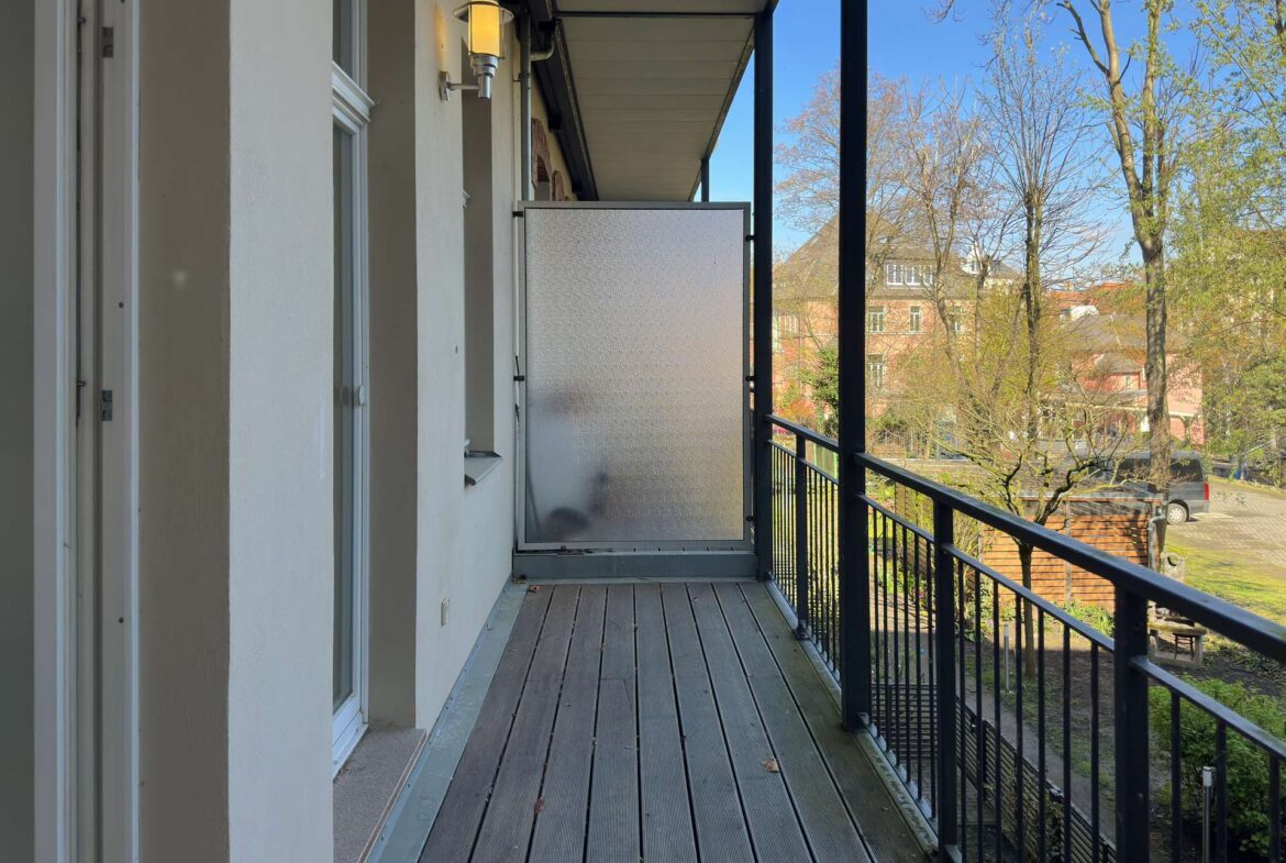 Narrow apartment balcony with wooden planks, frosted privacy screen at the far end, white walls, and black metal railing overlooking trees and buildings on a clear day.