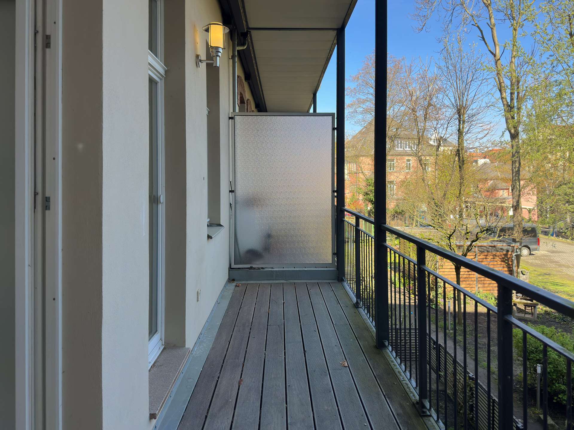 Narrow apartment balcony with wooden planks, frosted privacy screen at the far end, white walls, and black metal railing overlooking trees and buildings on a clear day.