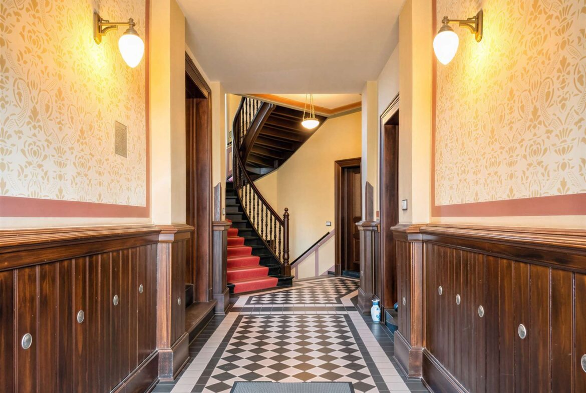 Elegant hotel hallway with a curved staircase, checkerboard floor and warm wall sconces on patterned wallpaper paneling.