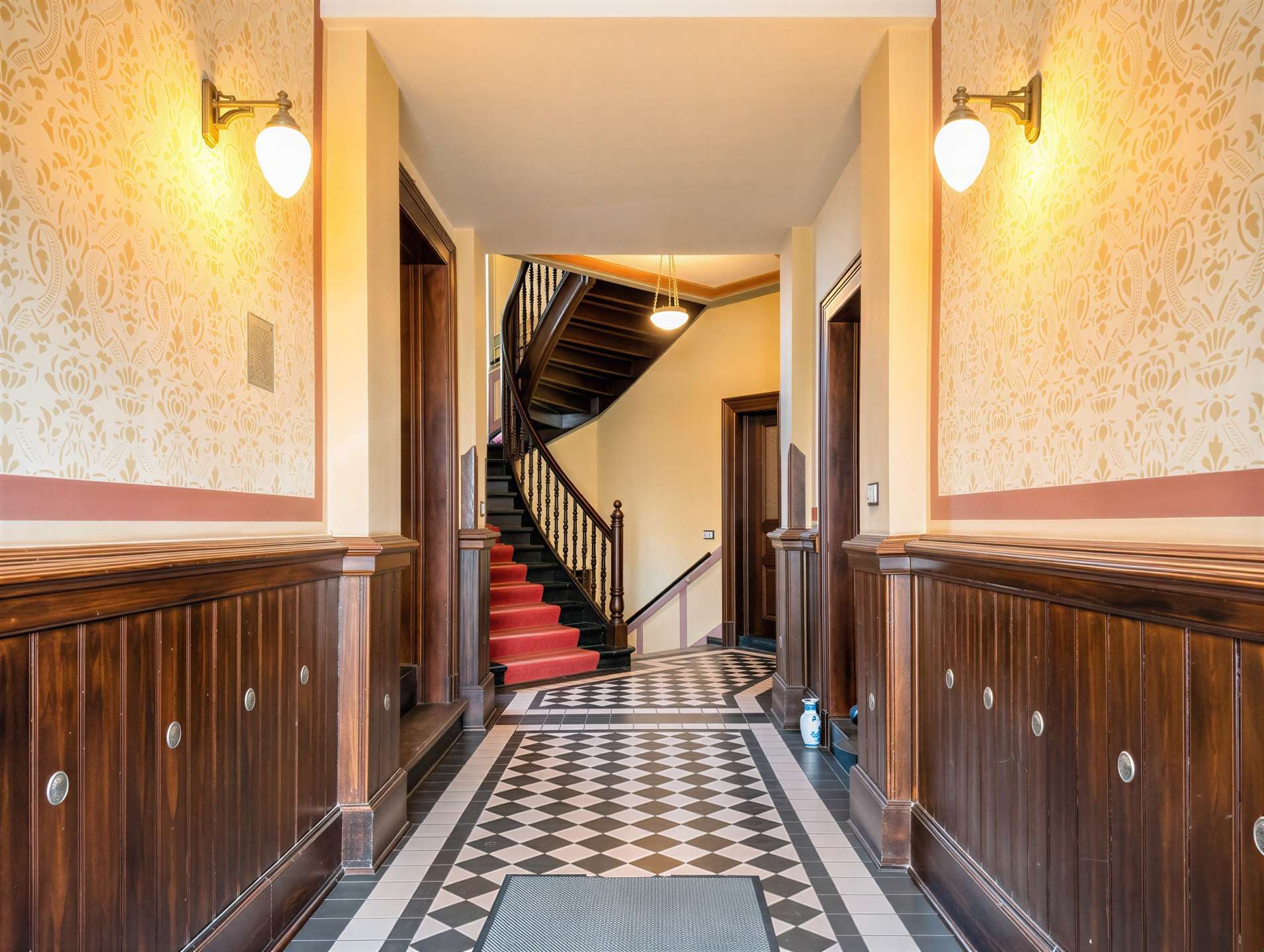 Elegant hotel hallway with a curved staircase, checkerboard floor and warm wall sconces on patterned wallpaper paneling.