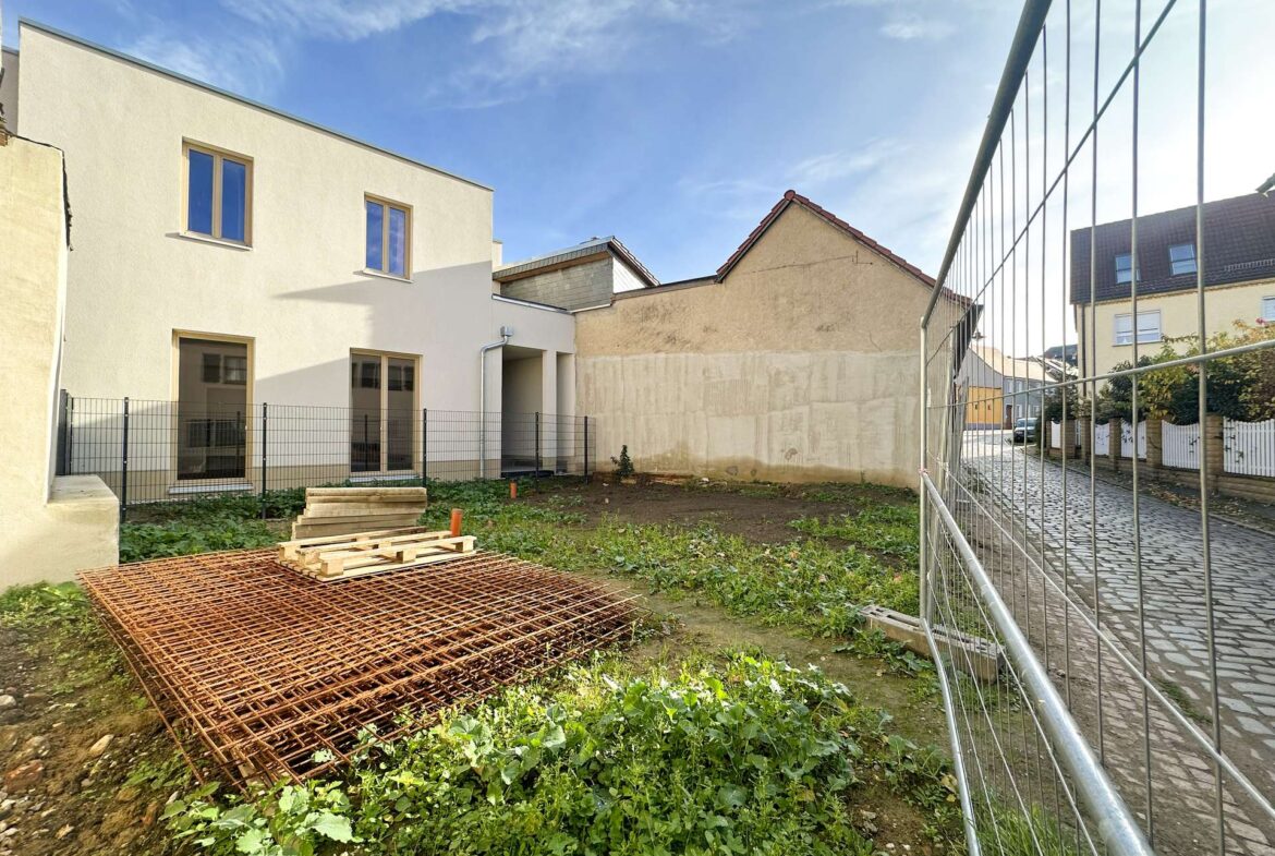 Construction yard with a metal fence, rusted rebar grid, and wooden pallets on bare soil near beige buildings under a blue sky.