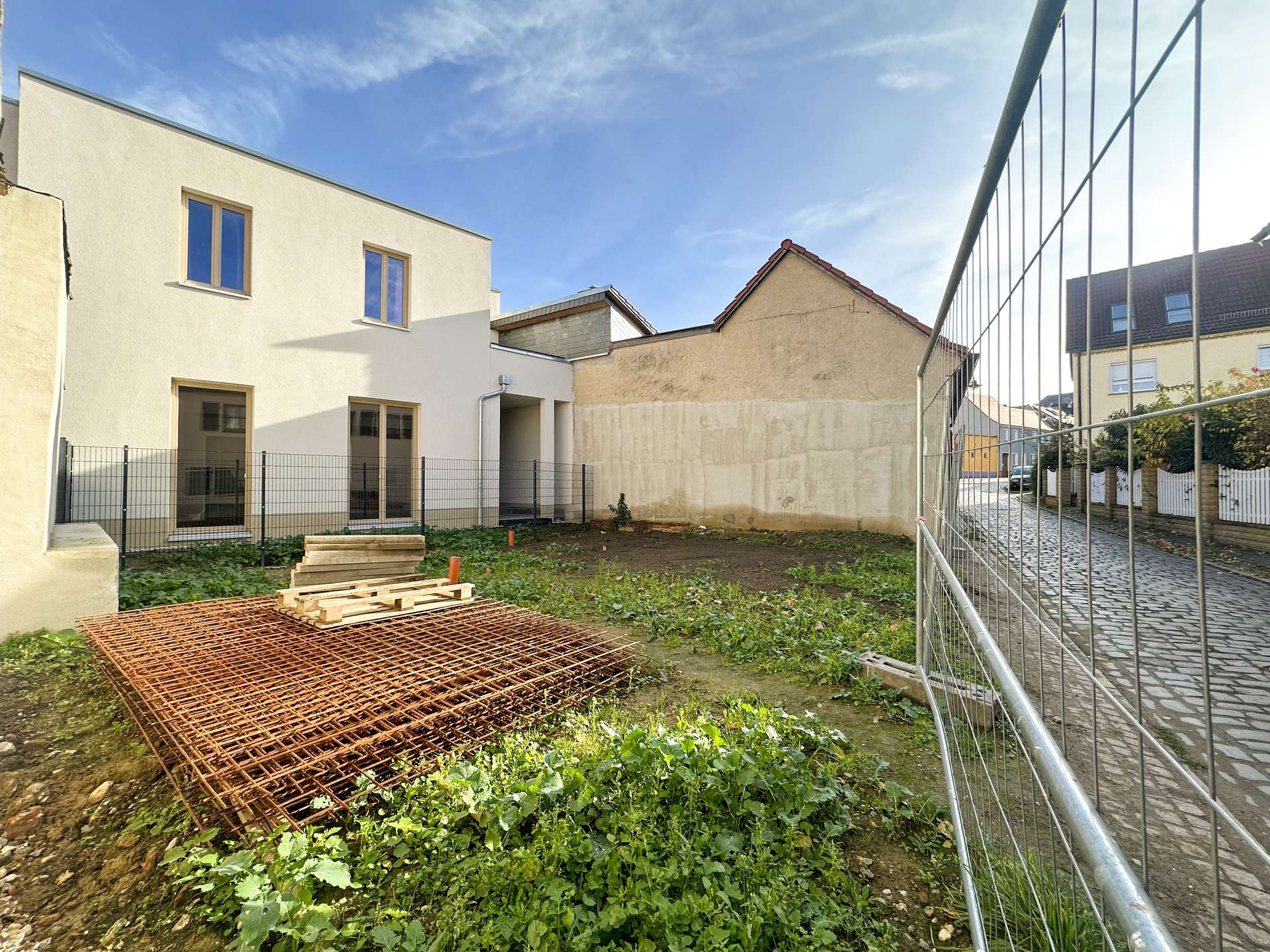 Construction yard with a metal fence, rusted rebar grid, and wooden pallets on bare soil near beige buildings under a blue sky.