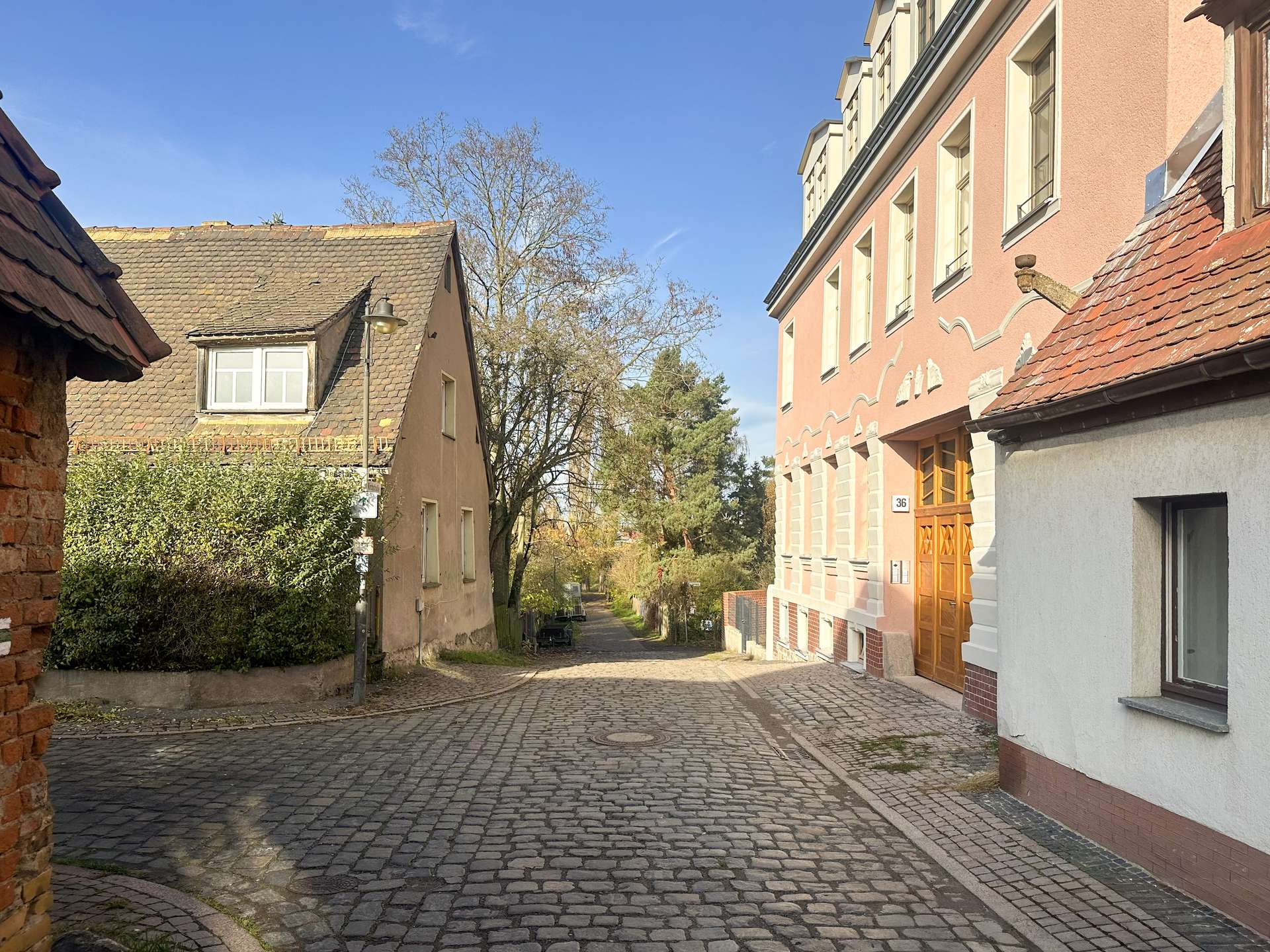 Cobblestone street between beige houses on the left and a pink decorative building on the right under a clear blue sky, in daylight.