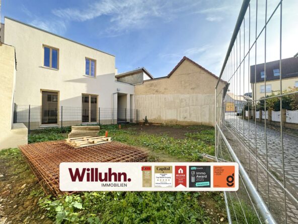 Construction site with a modern white building, metal fence, and a large Willuhn Immobilien sign showing awards.
