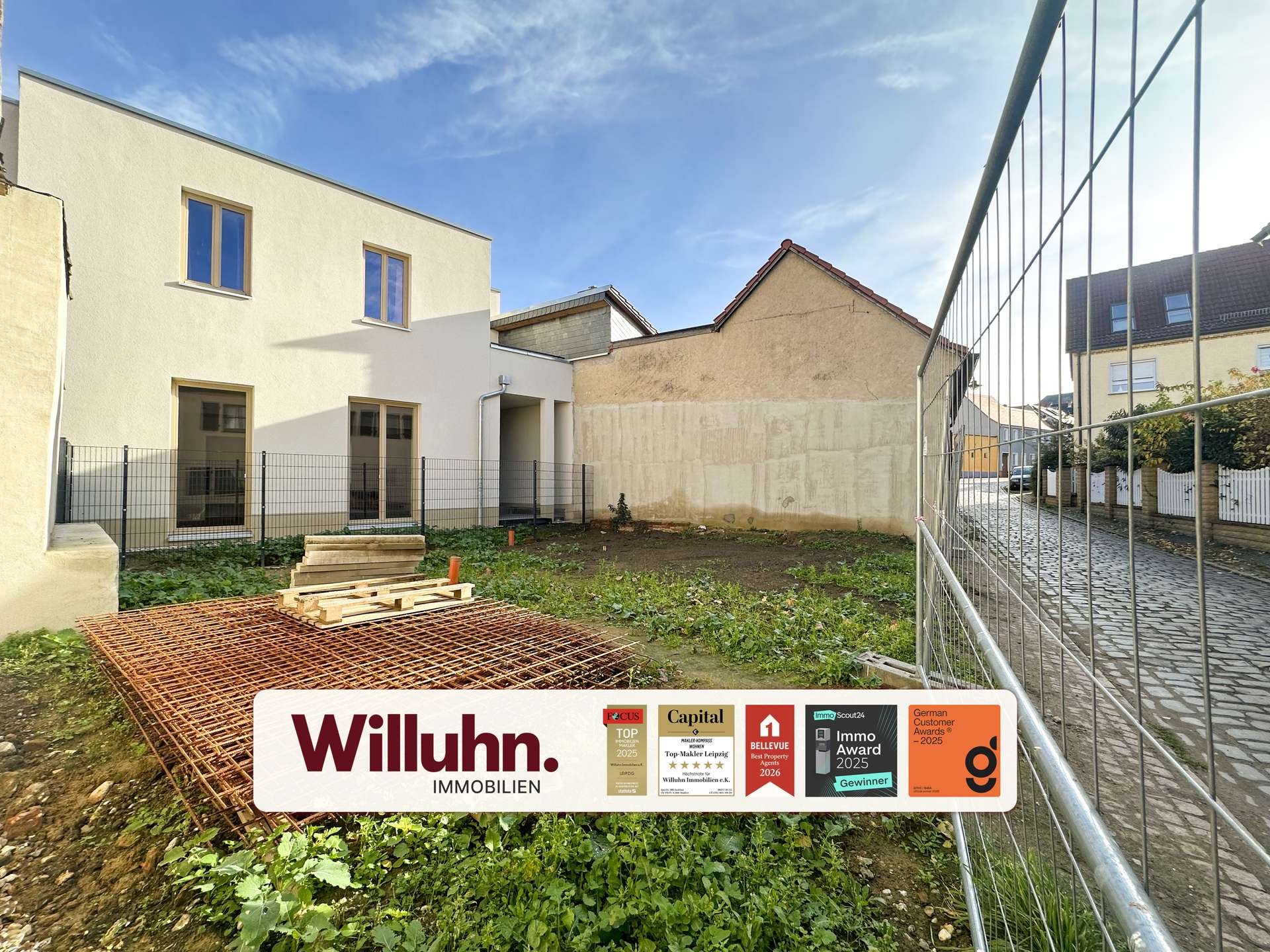 Construction site with a modern white building, metal fence, and a large Willuhn Immobilien sign showing awards.