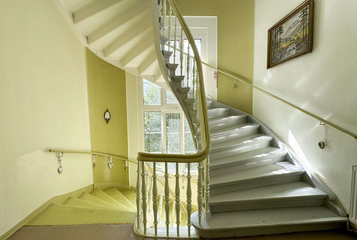 Curved white staircase with ornate railing in a pale yellow stairwell, a window at the landing, and a framed landscape painting on the wall to the right.