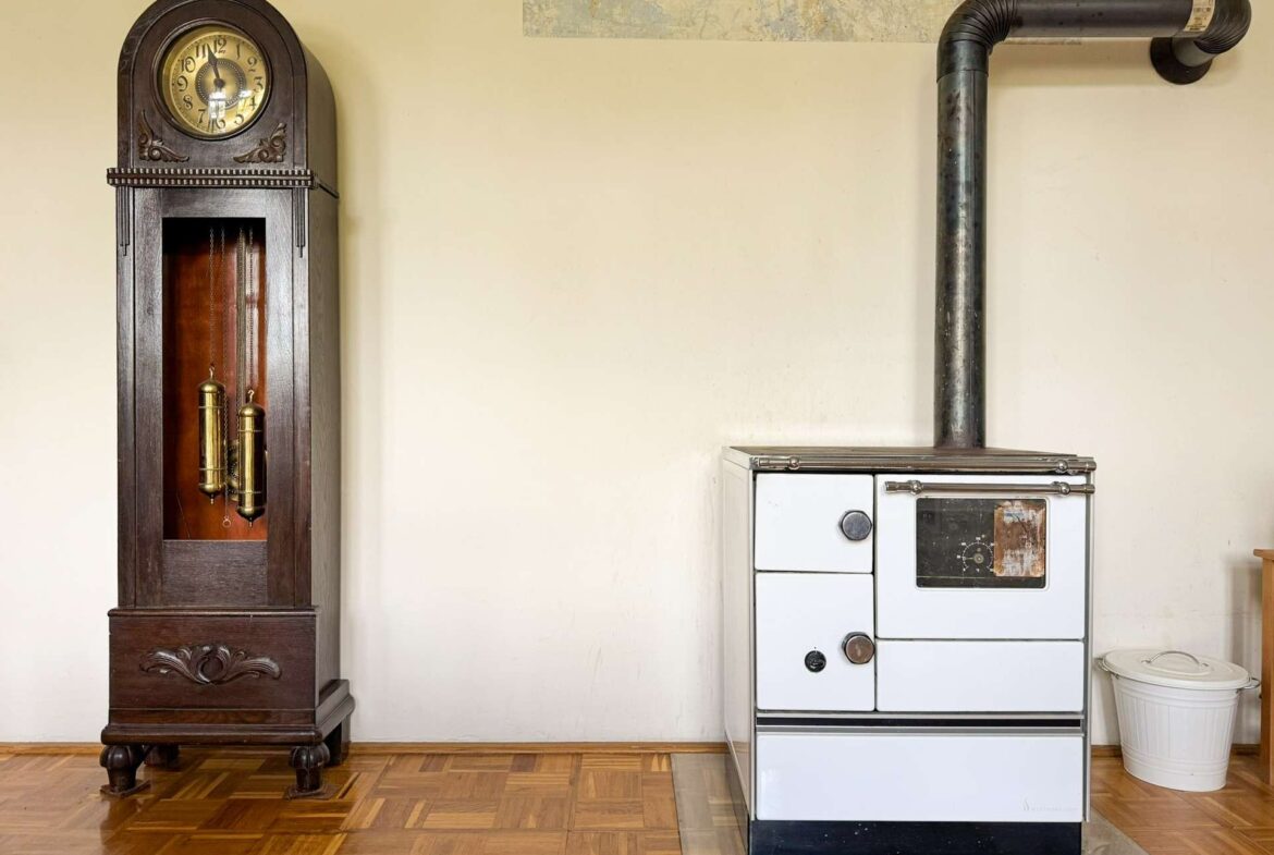 A tall, dark wooden grandfather clock next to an old white stove with a metal vent pipe against a beige wall in a vintage kitchen setting.
