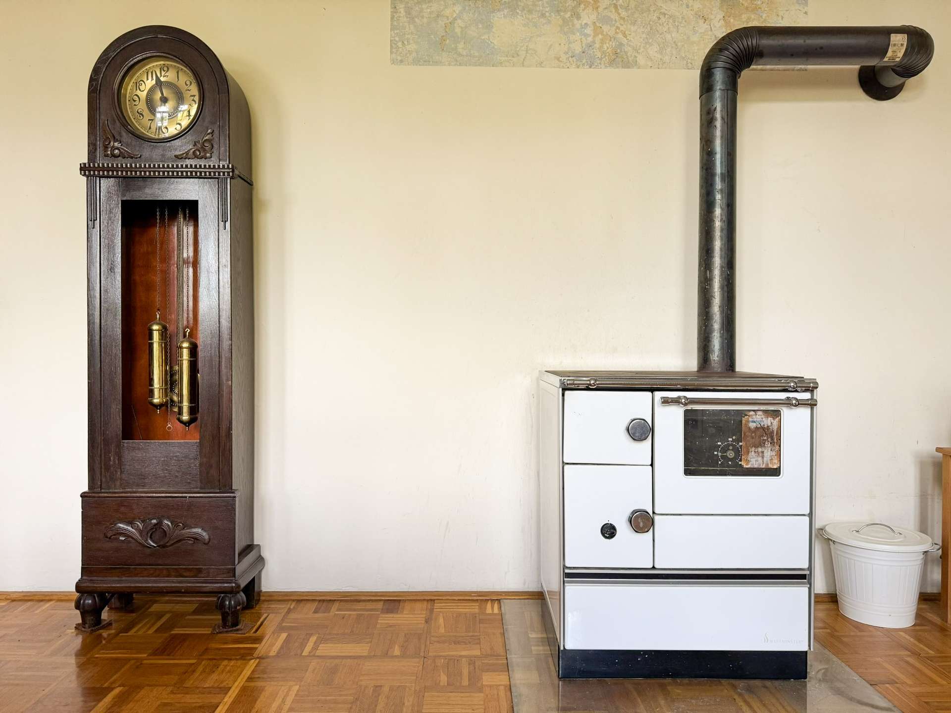 A tall, dark wooden grandfather clock next to an old white stove with a metal vent pipe against a beige wall in a vintage kitchen setting.