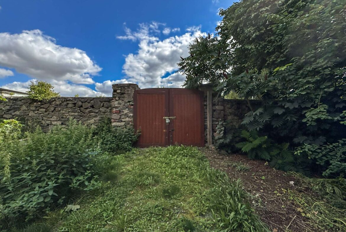 Red wooden gate set in a stone wall, surrounded by overgrown vegetation and blue sky above.