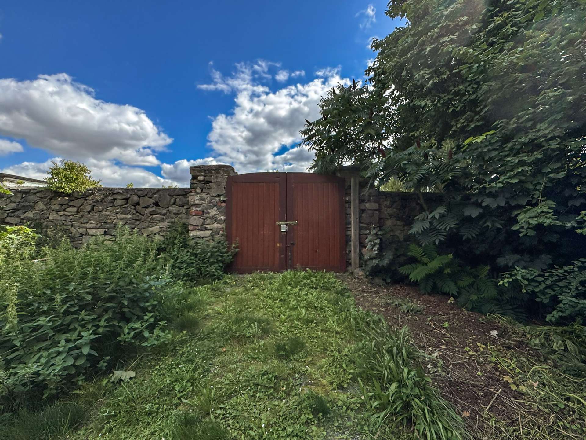 Red wooden gate set in a stone wall, surrounded by overgrown vegetation and blue sky above.
