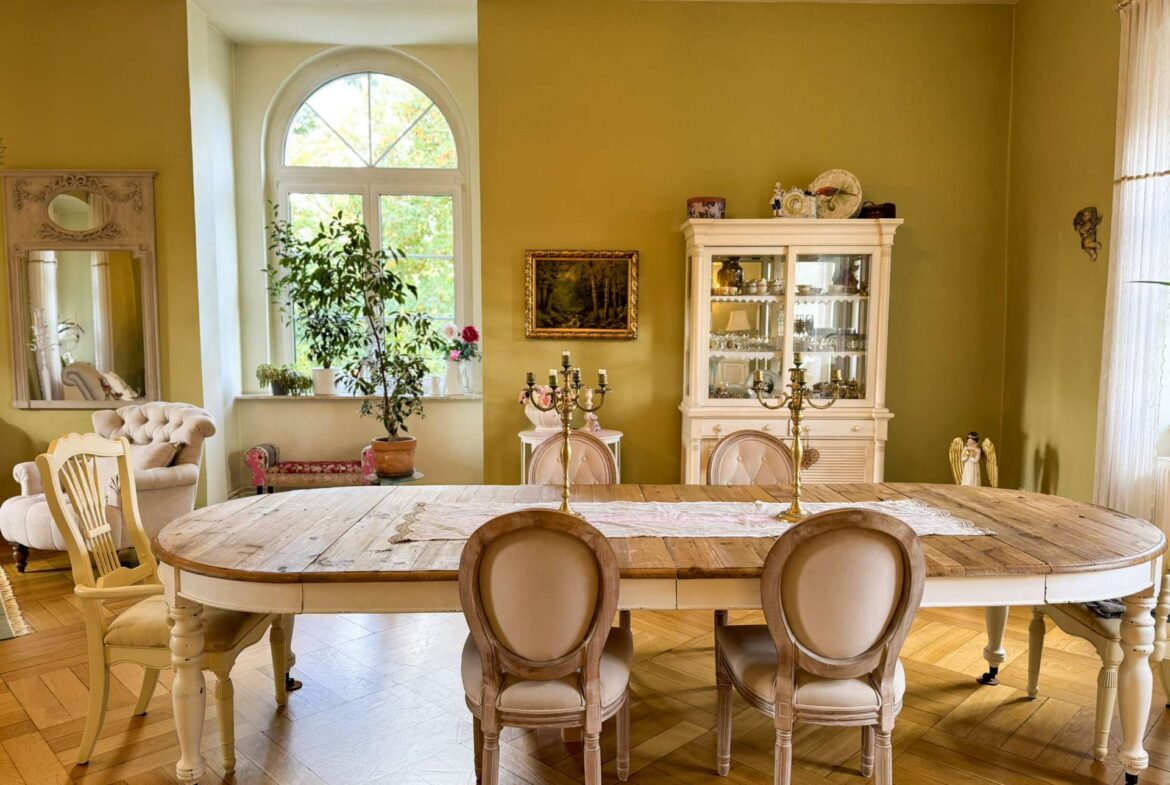 Vintage dining room with a large wooden table, cream upholstered chairs, and a glass-front china cabinet beside an arched window. A potted plant and decorative items adorn the room.