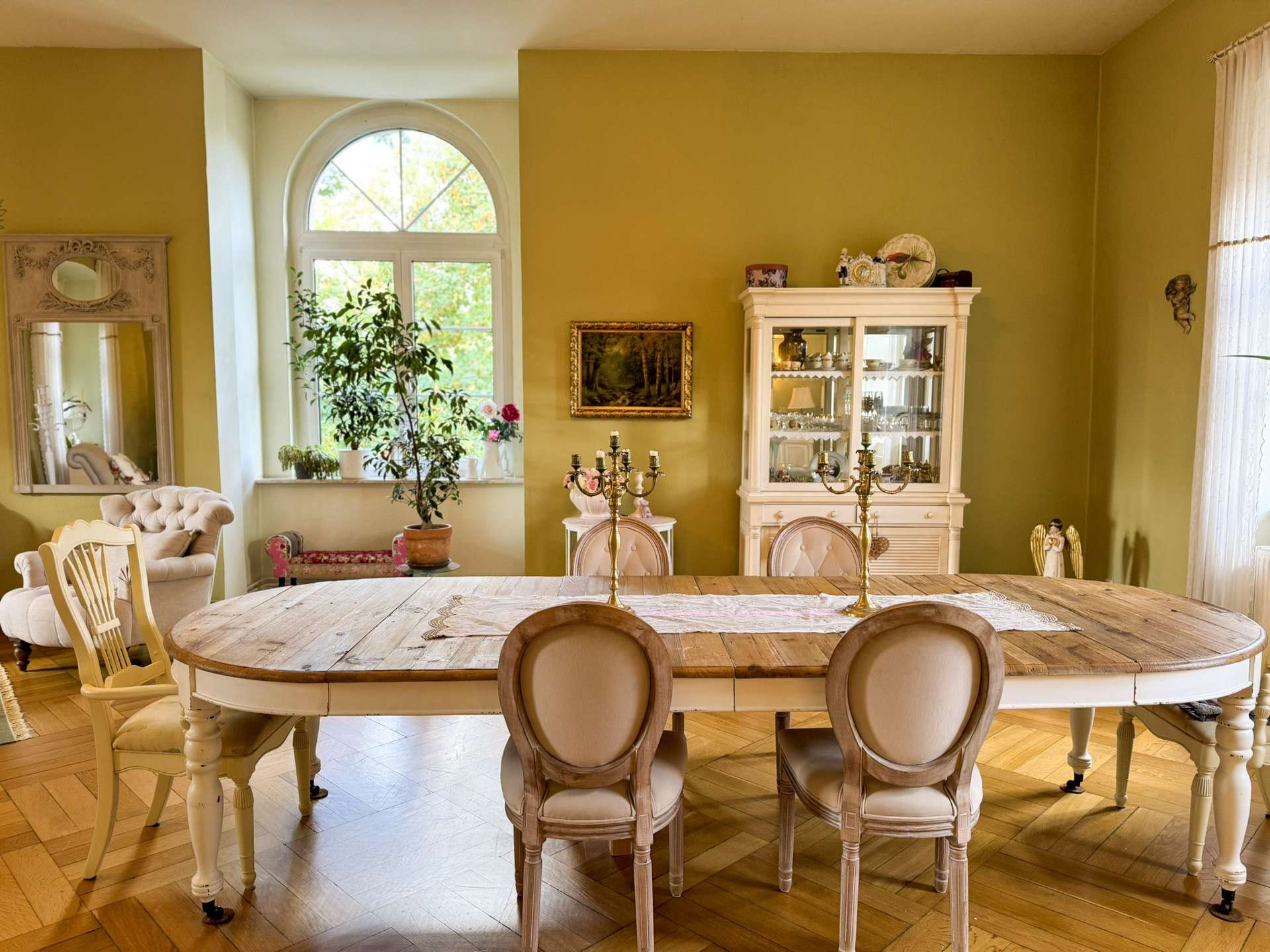 Vintage dining room with a large wooden table, cream upholstered chairs, and a glass-front china cabinet beside an arched window. A potted plant and decorative items adorn the room.