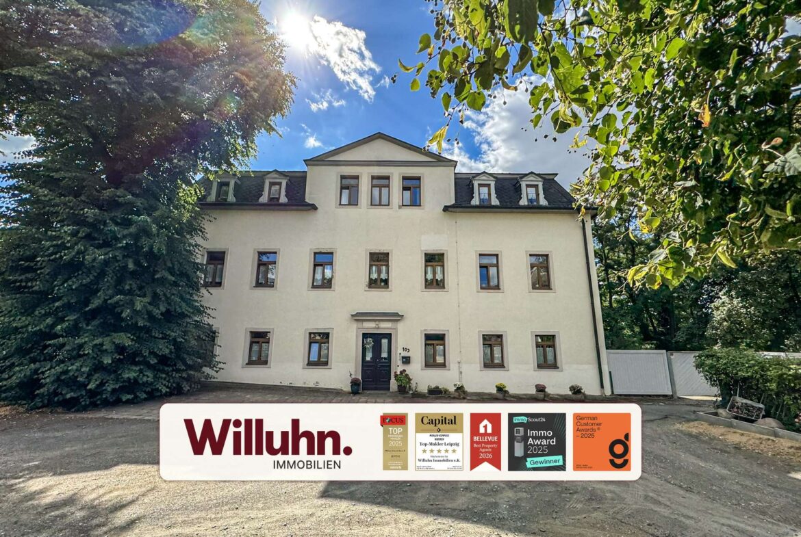 Front view of a pale multi-story building framed by green trees, with a large Willuhn Immobilien signboard displaying awards in the foreground.