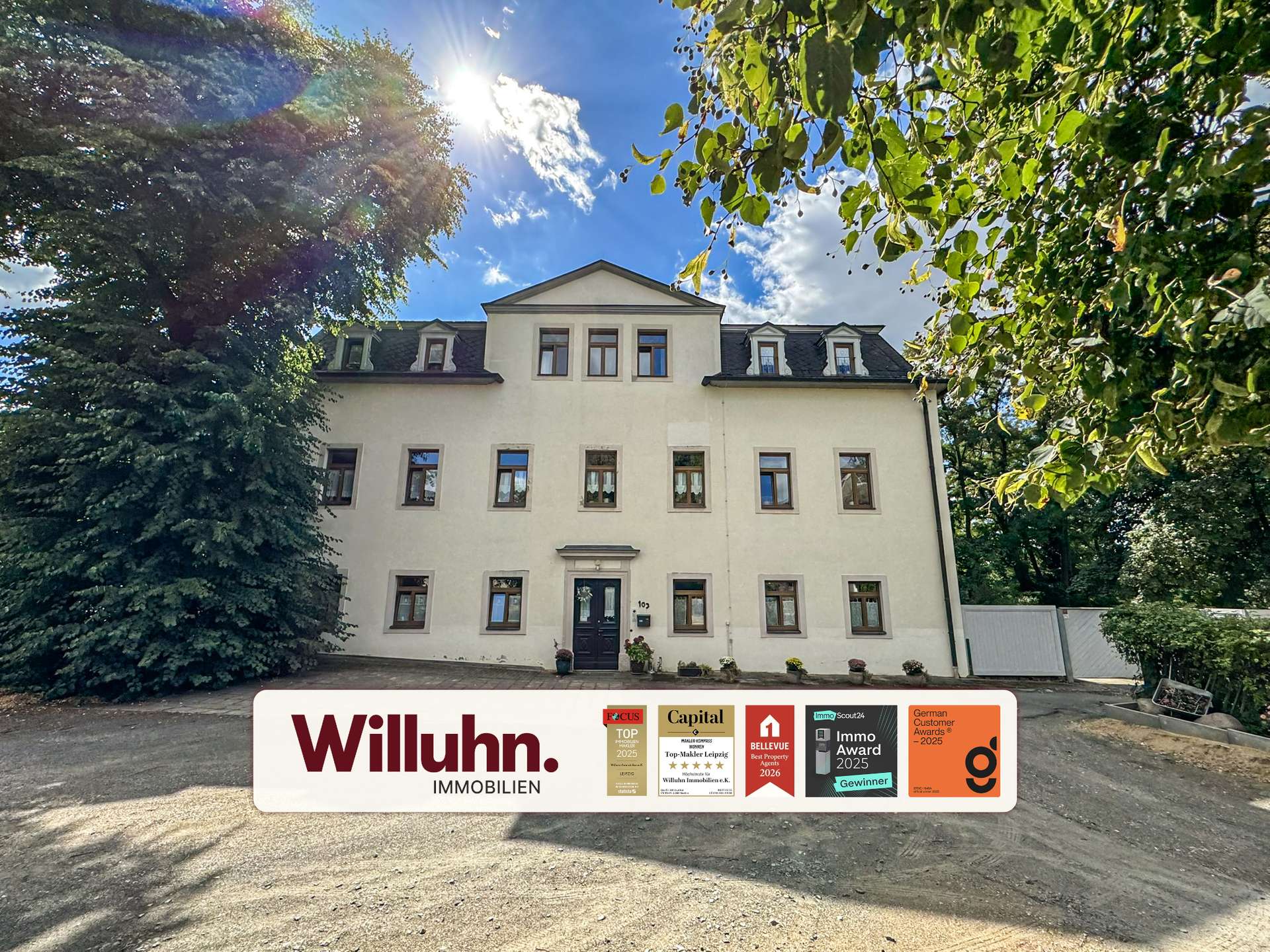 Front view of a pale multi-story building framed by green trees, with a large Willuhn Immobilien signboard displaying awards in the foreground.