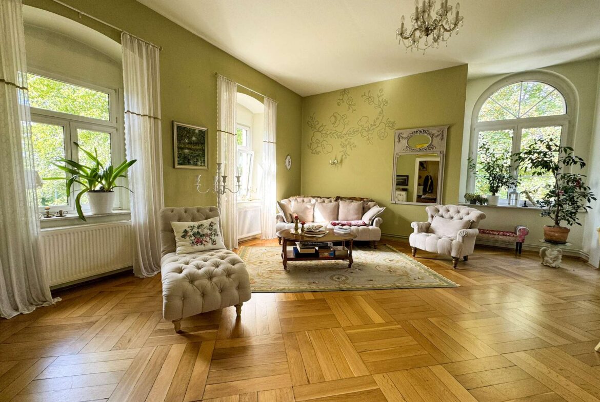 Light-filled living room with tufted beige sofas, large arched windows, and a chandelier; pale green walls with floral wall art and potted plants along the windowsill.
