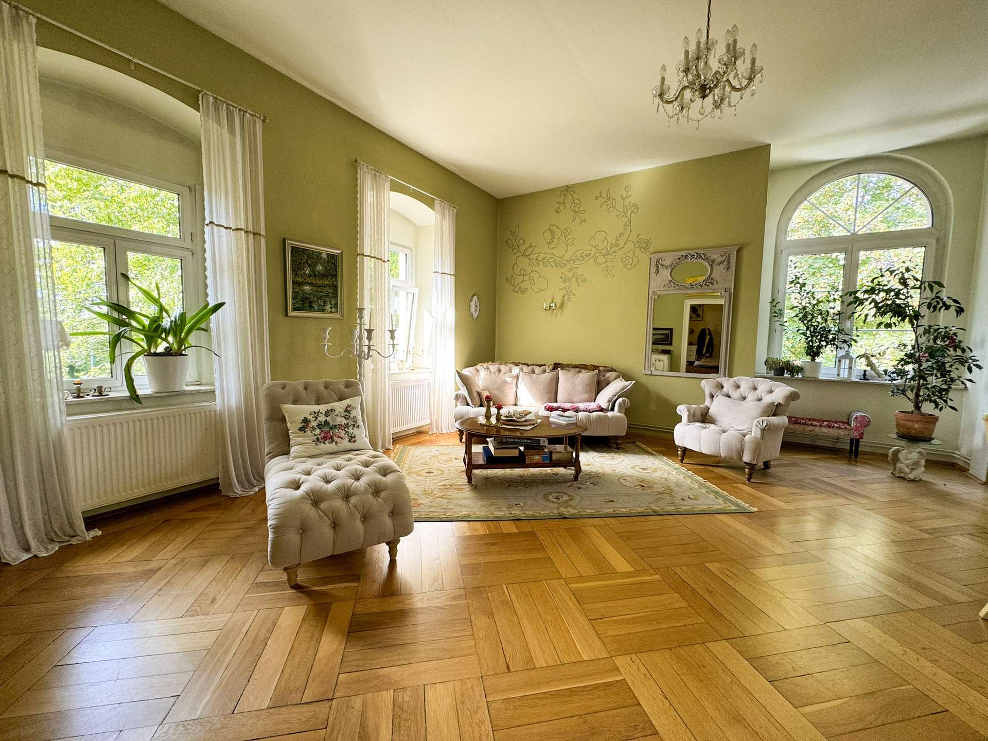Light-filled living room with tufted beige sofas, large arched windows, and a chandelier; pale green walls with floral wall art and potted plants along the windowsill.