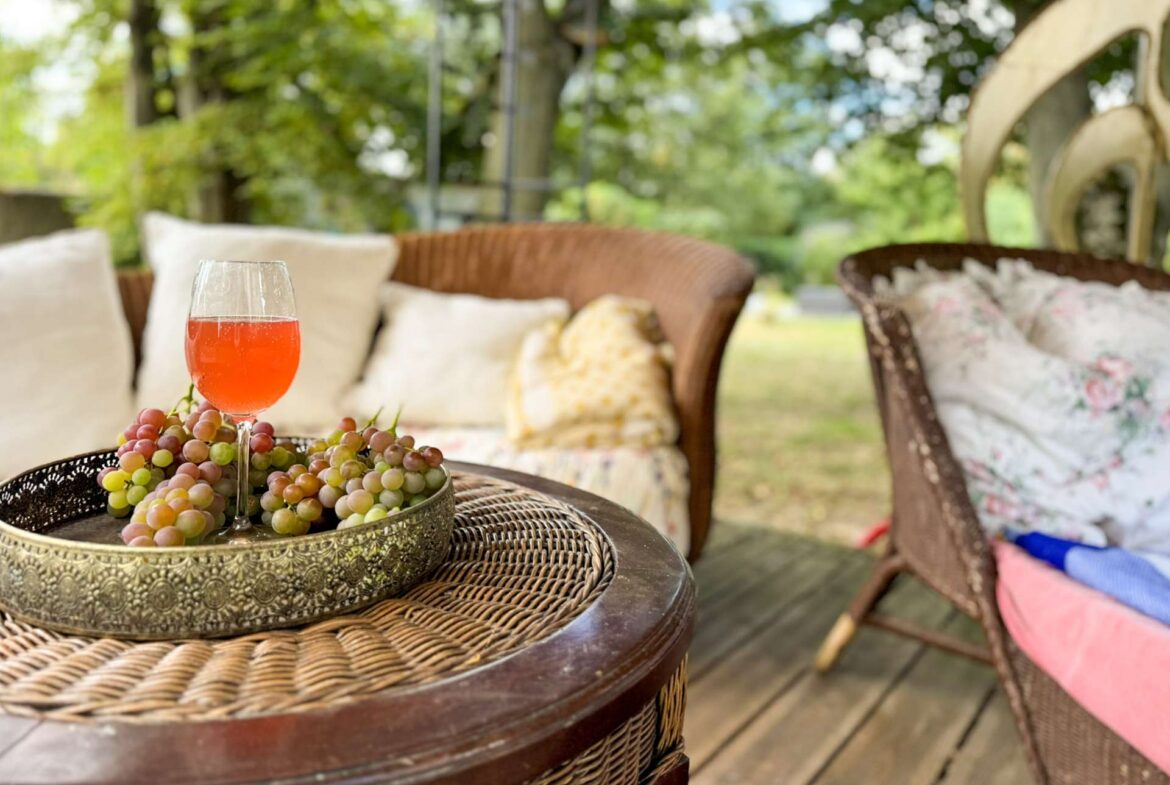Glass of pink beverage and a bunch of grapes on a decorative metal tray on a round wicker table in a sunny garden lounge.
