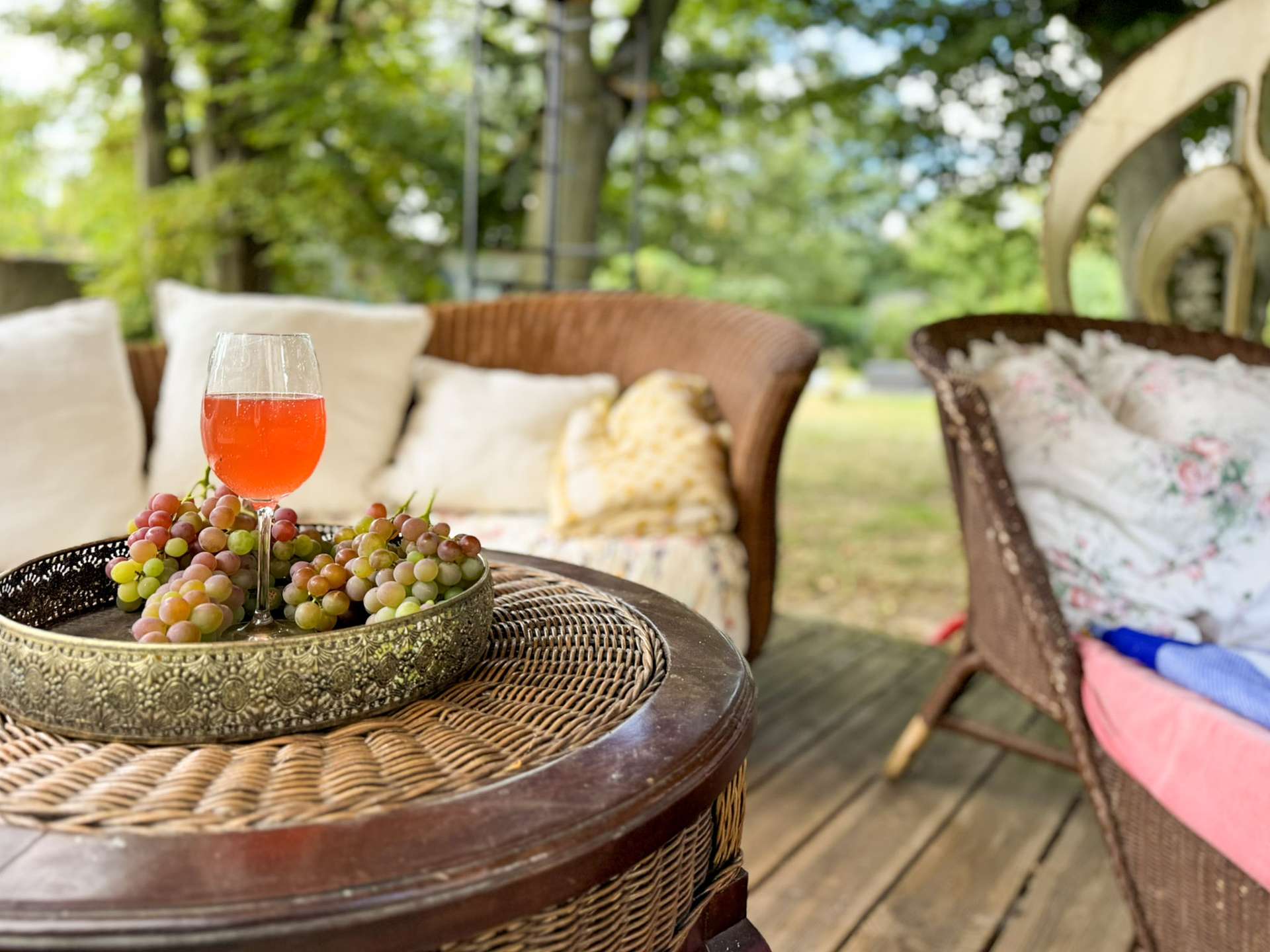 Glass of pink beverage and a bunch of grapes on a decorative metal tray on a round wicker table in a sunny garden lounge.