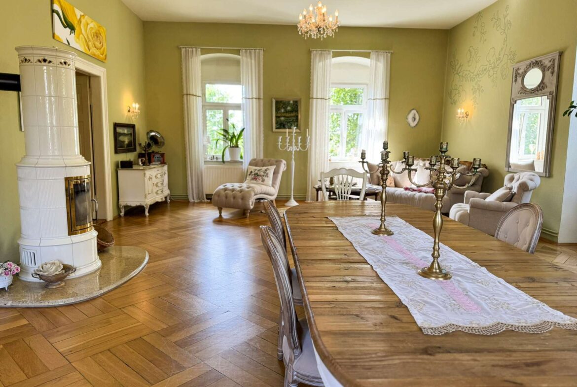 Bright living and dining room with green walls, a long wooden dining table dressed with a white lace runner and brass candelabras, and a white tiled stove on the left.