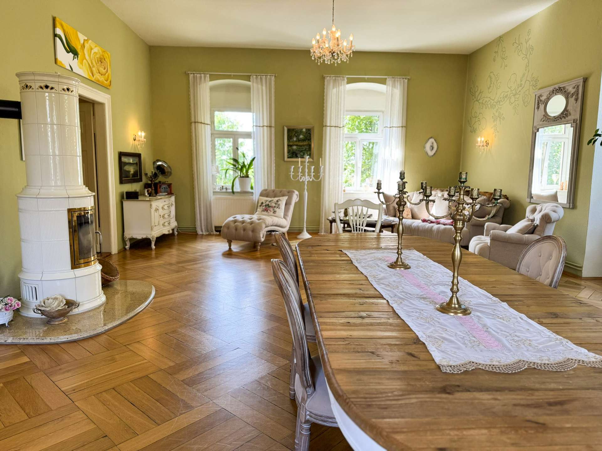 Bright living and dining room with green walls, a long wooden dining table dressed with a white lace runner and brass candelabras, and a white tiled stove on the left.