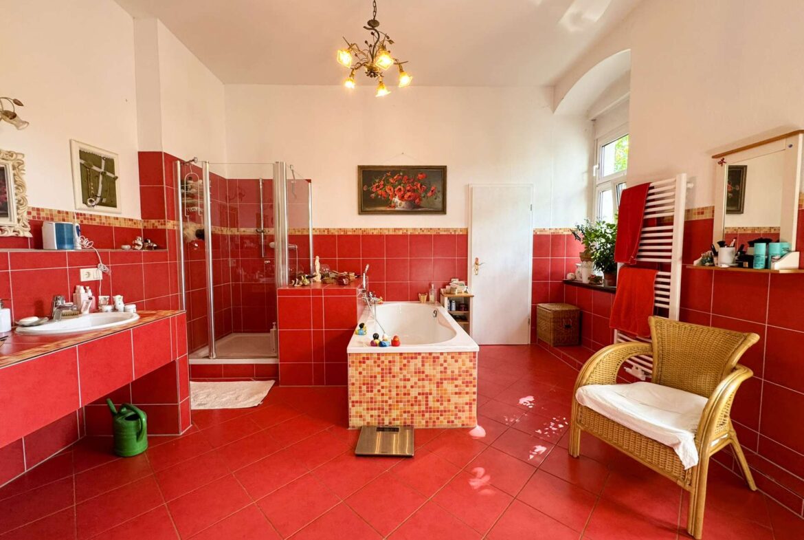 Bright red-tiled bathroom featuring a corner glass shower, freestanding tub, and a wicker chair near a window with potted plants; towels adorn a white heated rack on the right.
