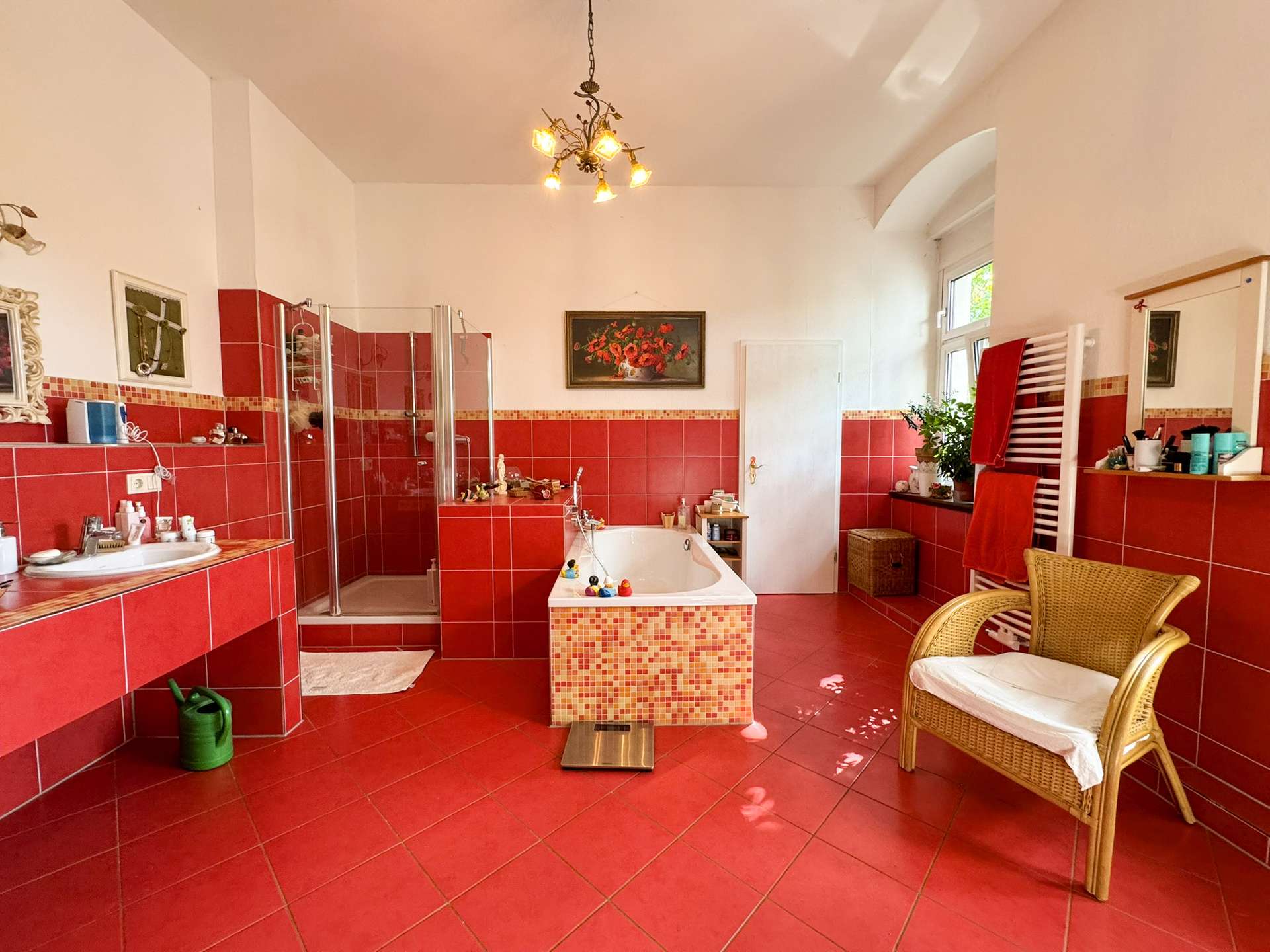 Bright red-tiled bathroom featuring a corner glass shower, freestanding tub, and a wicker chair near a window with potted plants; towels adorn a white heated rack on the right.