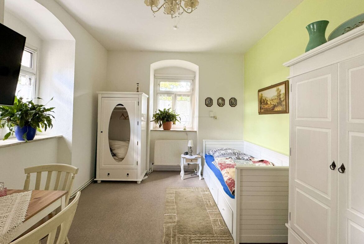 Light-filled vintage bedroom with white wardrobes, a single bed, dresser, and potted plants.