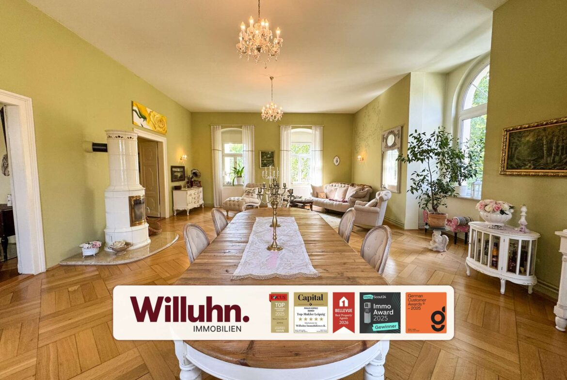 Bright living and dining room with green walls, a long wooden table, cream chairs, and an ornate chandelier; plants and arched windows in back. A Willuhn Immobilien banner with award badges is at the bottom.
