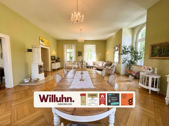 Bright living and dining room with green walls, a long wooden table, cream chairs, and an ornate chandelier; plants and arched windows in back. A Willuhn Immobilien banner with award badges is at the bottom.