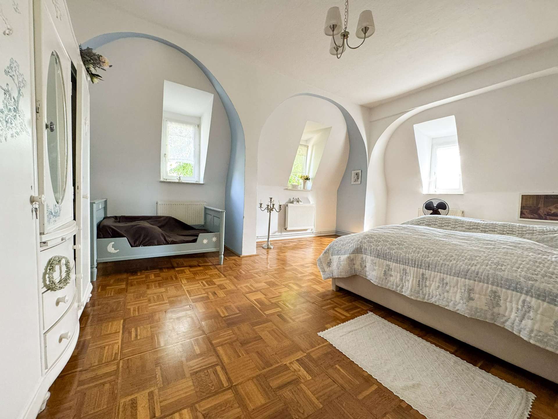 Bright bedroom with curved arches, a large bed on the right, a blue daybed on the left, and a chandelier overhead.