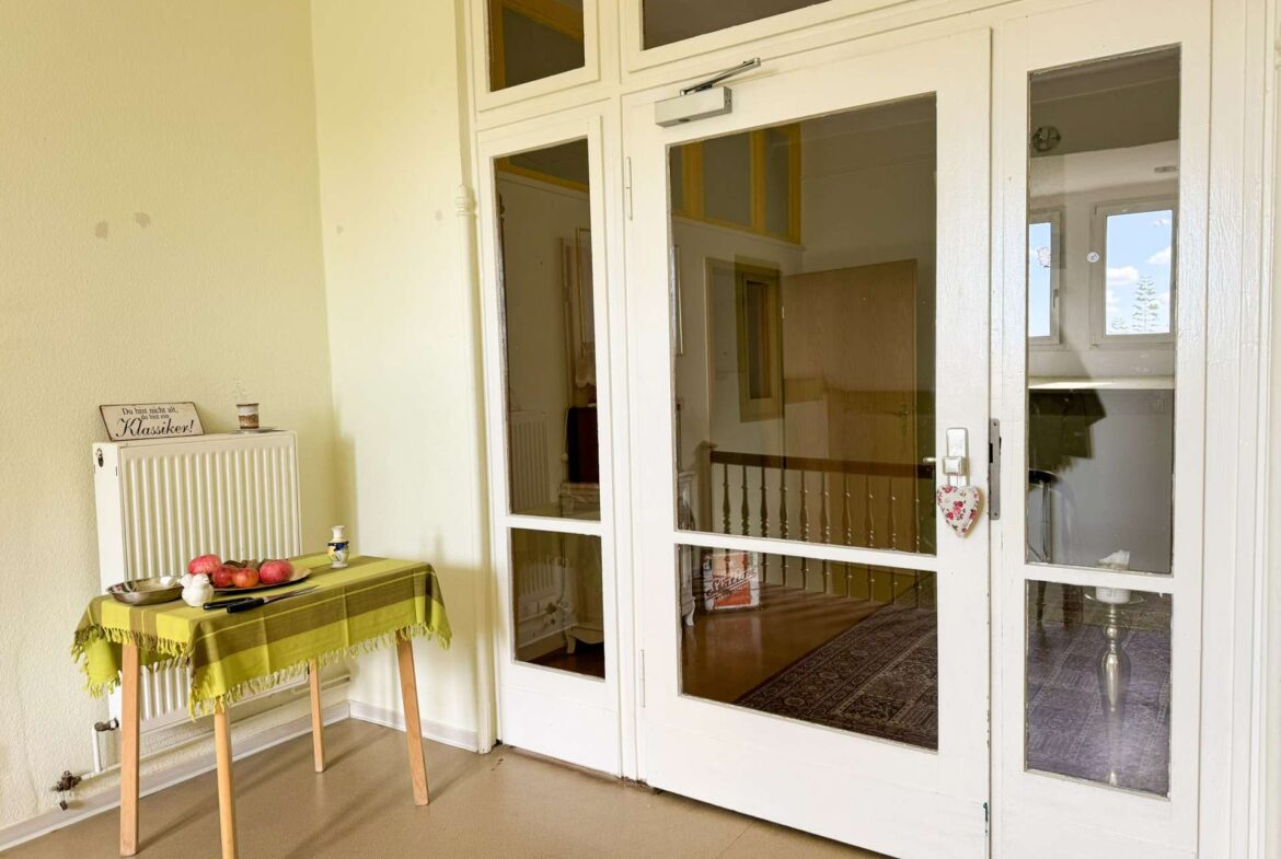 Interior scene near glass doors with a small table covered by a yellow-green tablecloth and fruit; a radiator on the left and a decorative sign above it reads 'Du bist nicht alt, du bist ein Klassiker!'