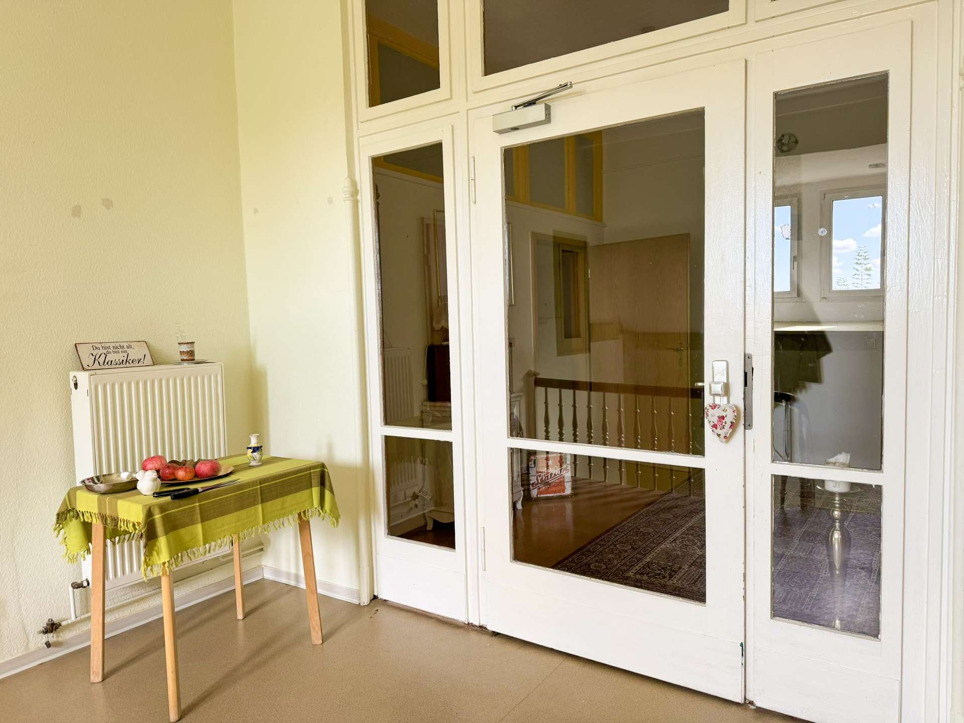 Interior scene near glass doors with a small table covered by a yellow-green tablecloth and fruit; a radiator on the left and a decorative sign above it reads 'Du bist nicht alt, du bist ein Klassiker!'
