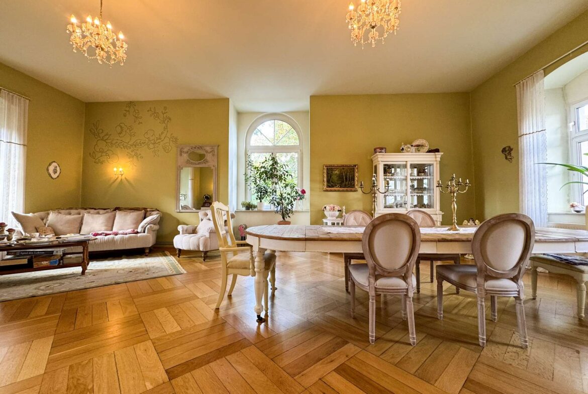 Elegant dining room and living area with a large oval table, cream chairs, chandelier, and china cabinet; warm yellow walls and parquet floor.