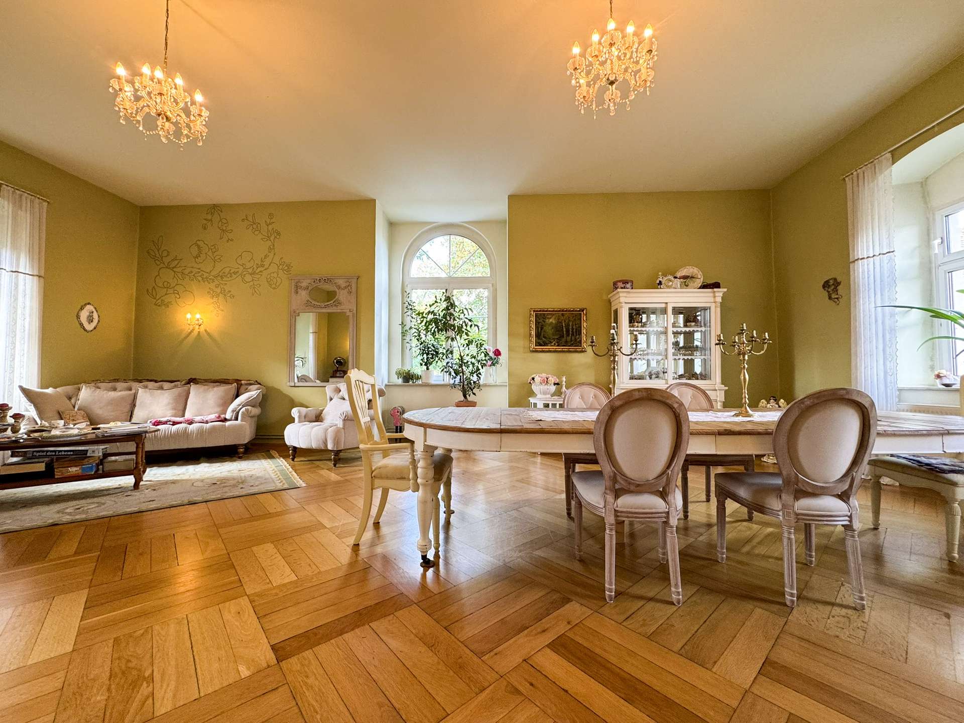 Elegant dining room and living area with a large oval table, cream chairs, chandelier, and china cabinet; warm yellow walls and parquet floor.