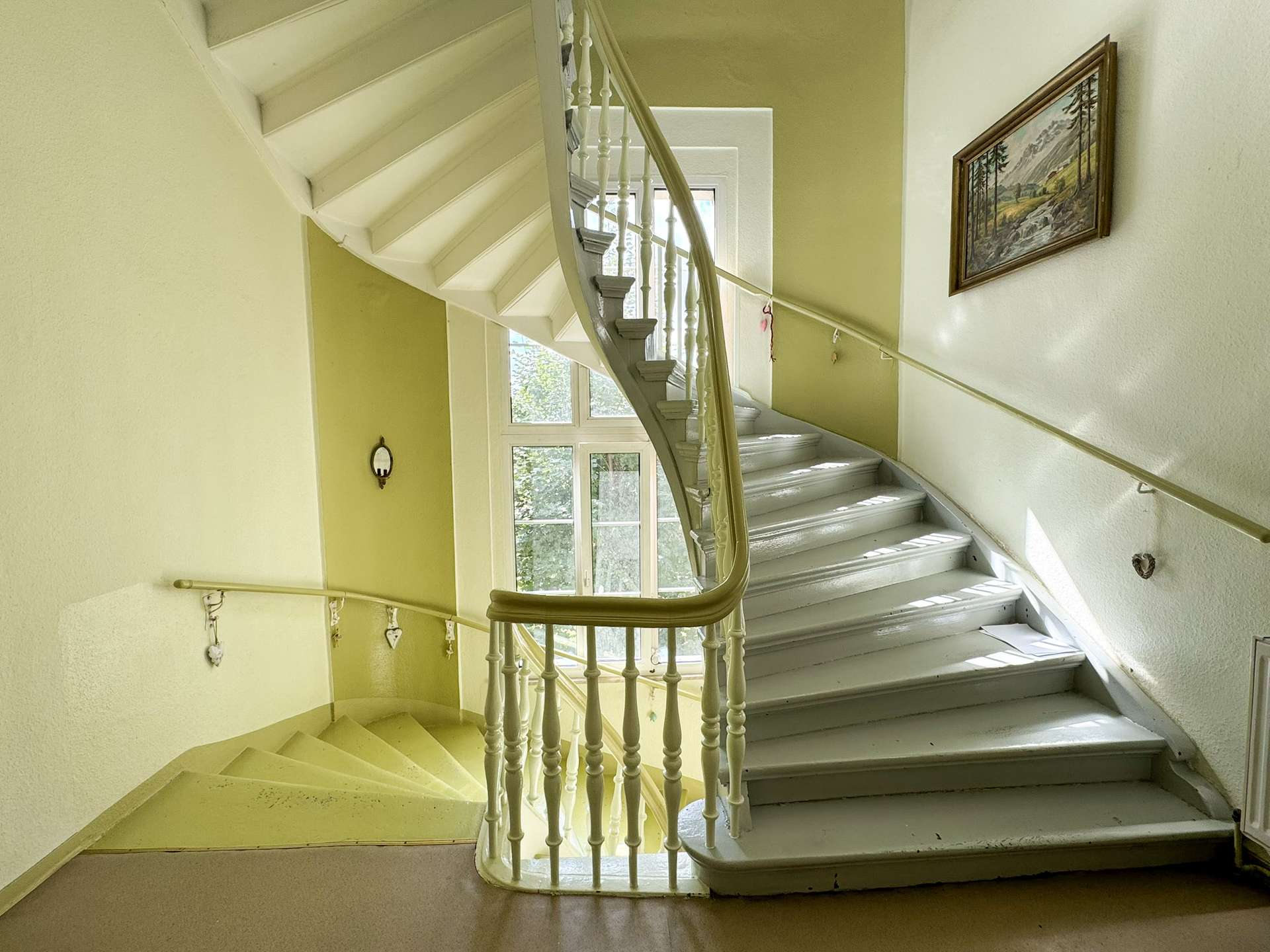 Curved white staircase with decorative balusters in a sunlit yellow stairwell, window at the landing, and a framed landscape painting on the wall.