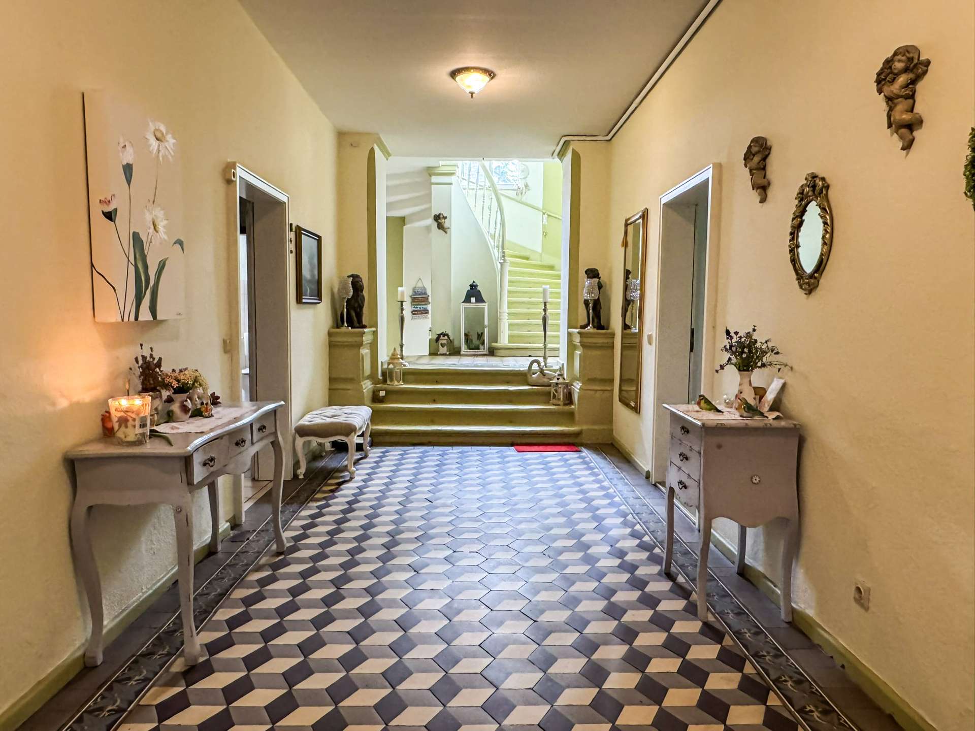 Long beige hallway with a blue-and-white checkered tiled floor, console tables with candles and vases, and a curved staircase at the far end.