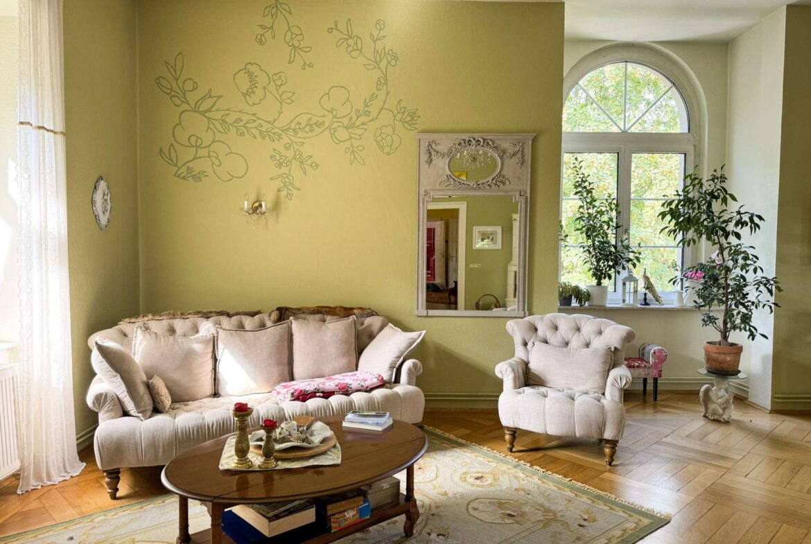 Elegant living room with olive walls, a large floral wall mural, cream tufted sofas, and a wooden coffee table by a big arched window.