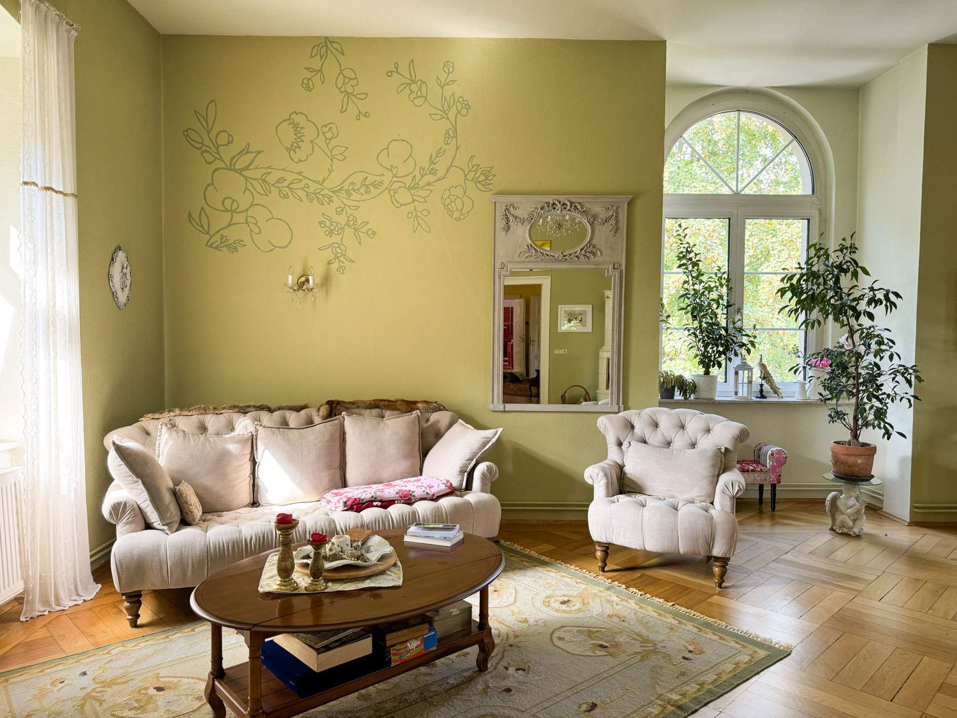 Elegant living room with olive walls, a large floral wall mural, cream tufted sofas, and a wooden coffee table by a big arched window.